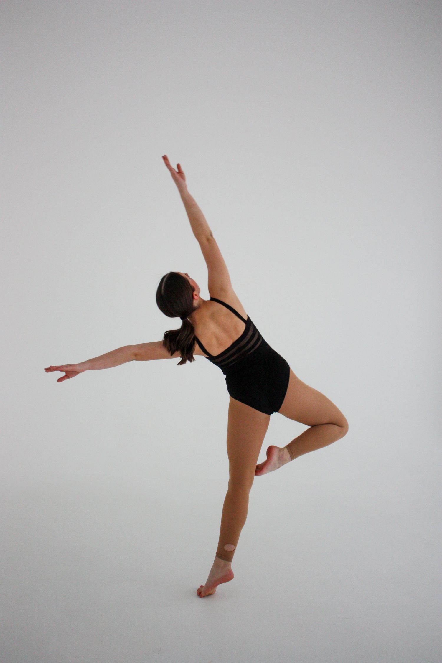 A female dancer in a black leotard balances on one leg with arm raised in an elegant studio pose.
