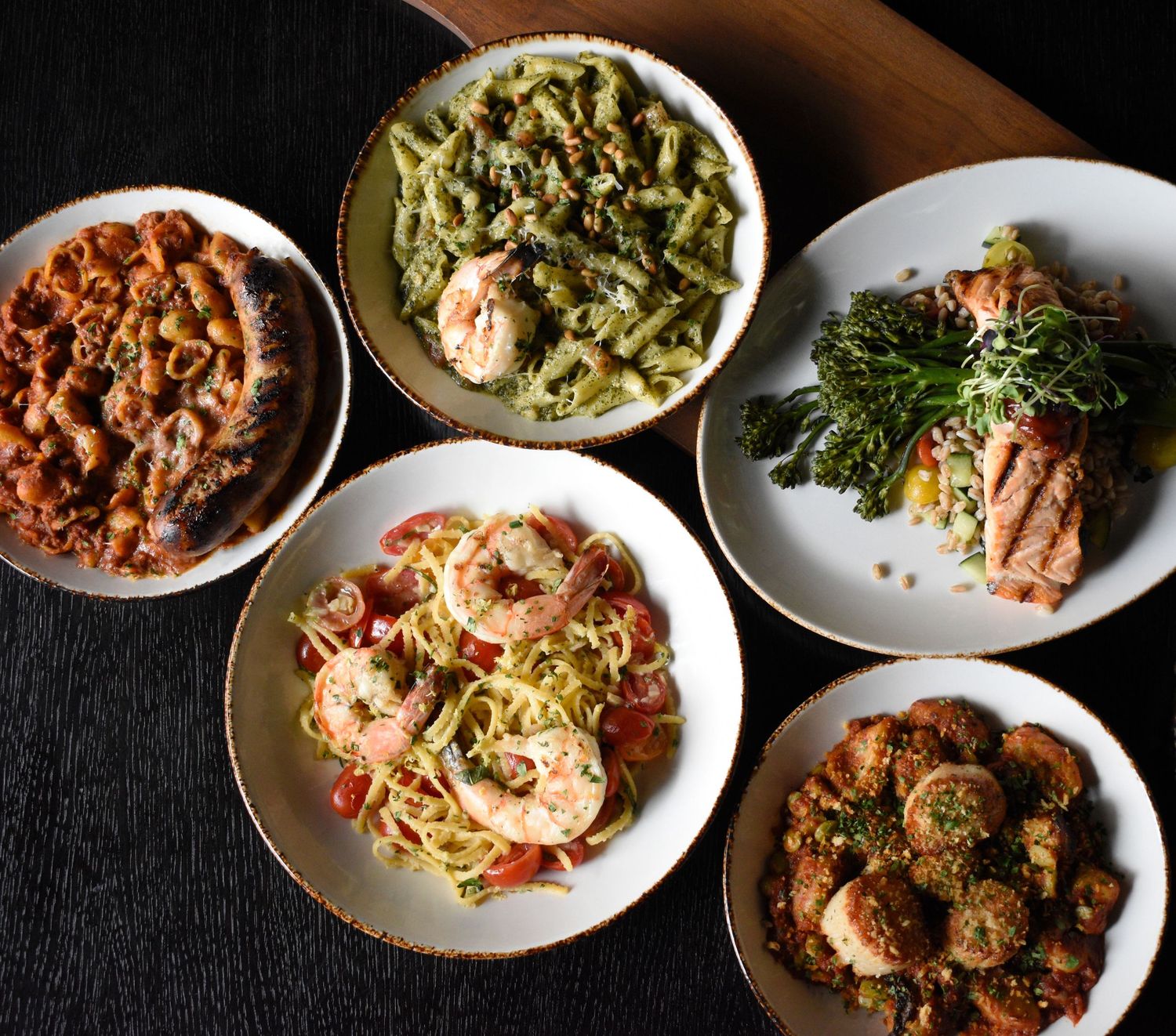 Overhead view of five Italian pasta and seafood dishes arranged on a dark wooden table.