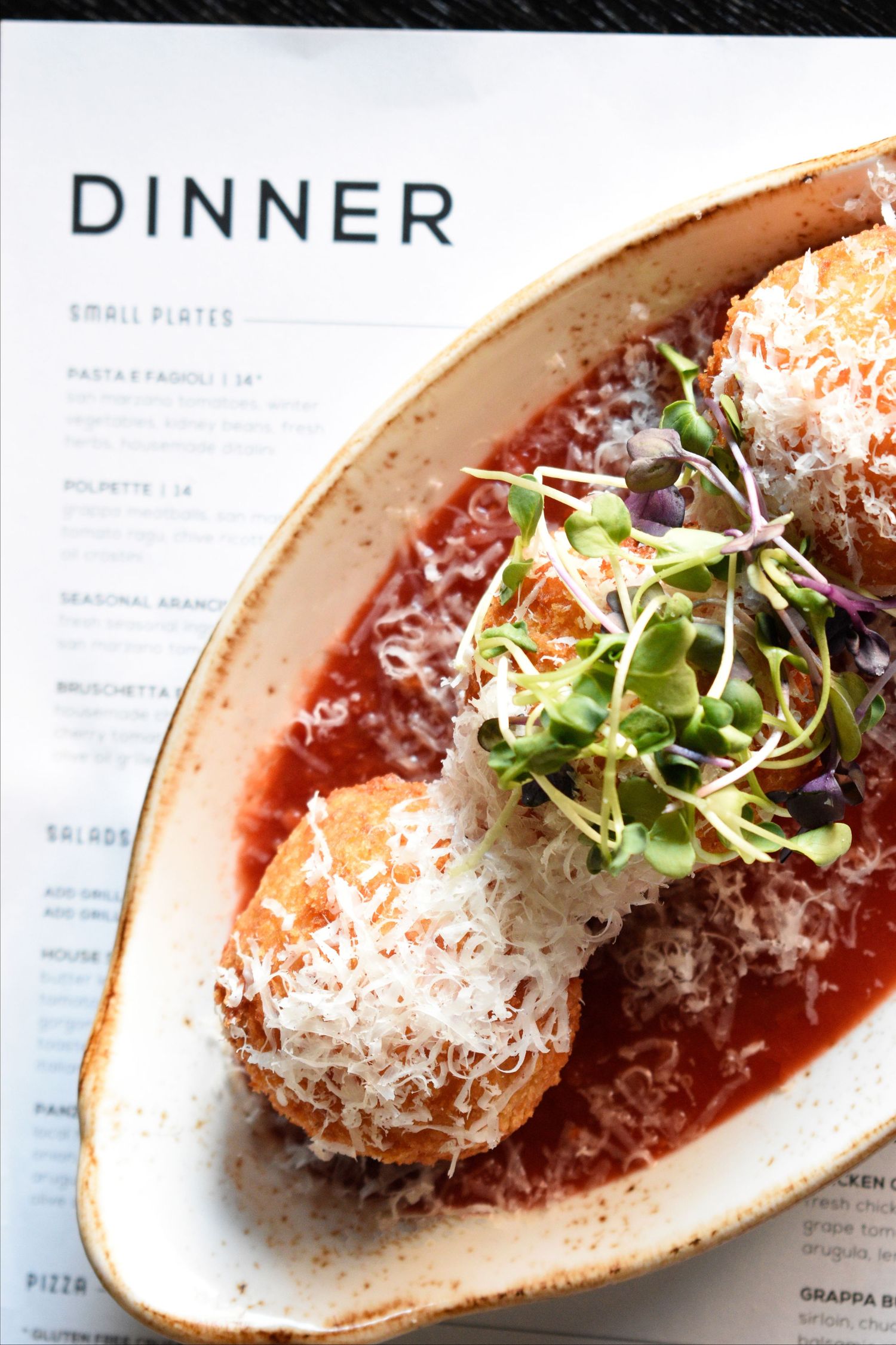 Restaurant arancini dish in tomato sauce with parmesan and microgreens next to an Italian dinner menu.