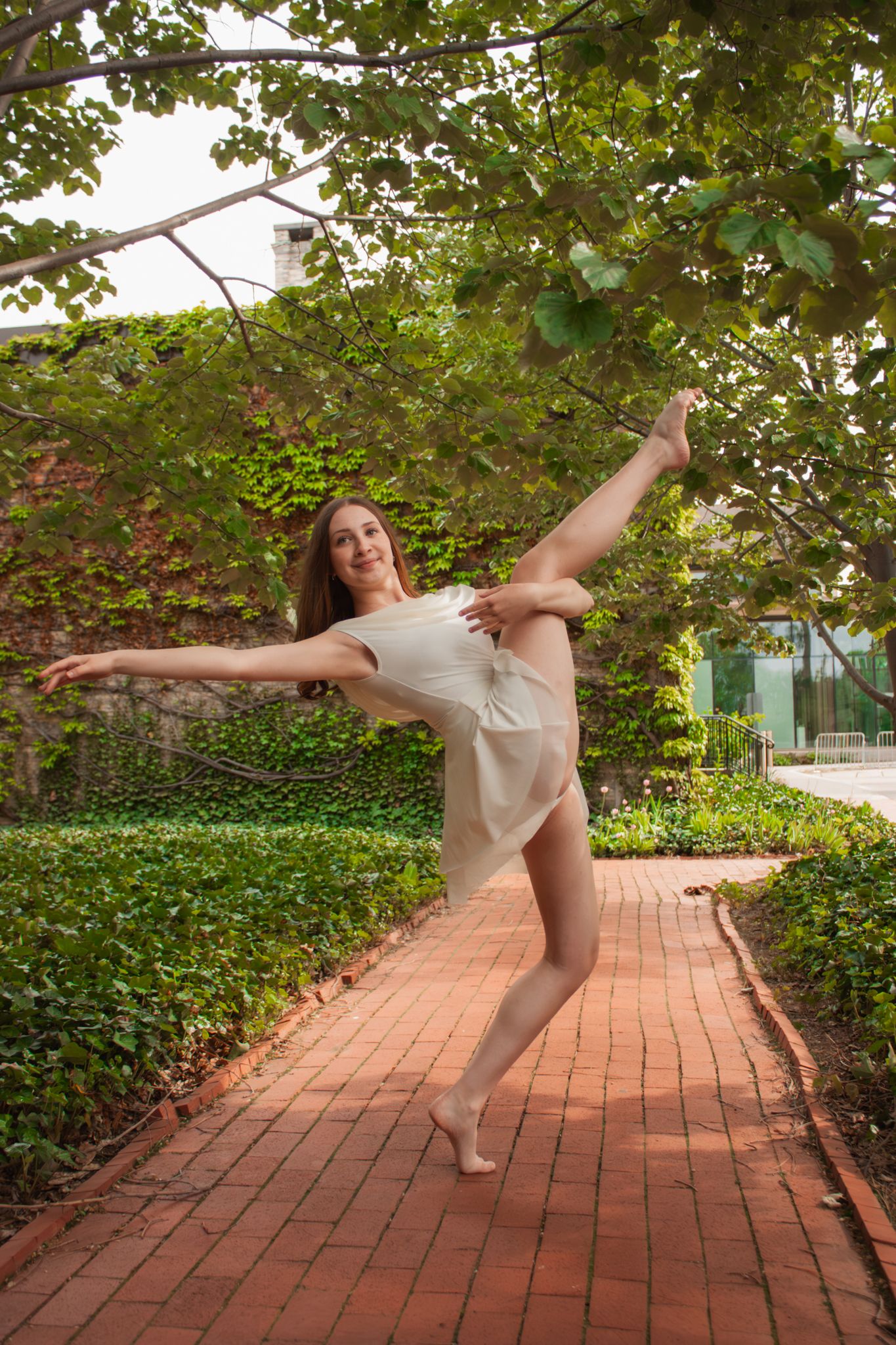 A graceful ballet dancer performs an arabesque on a brick path surrounded by lush green ivy and trees.