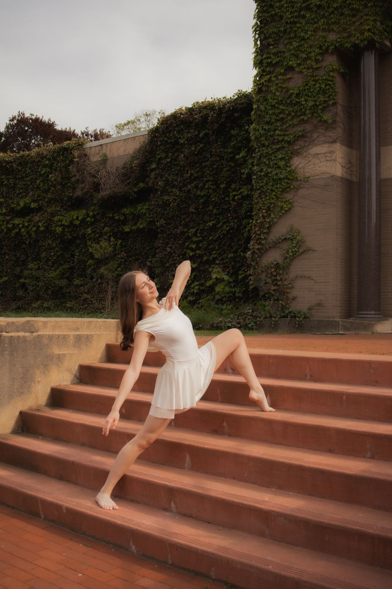 A graceful dancer in a white dress poses elegantly on brick steps beside an ivy-covered stone building outdoors.