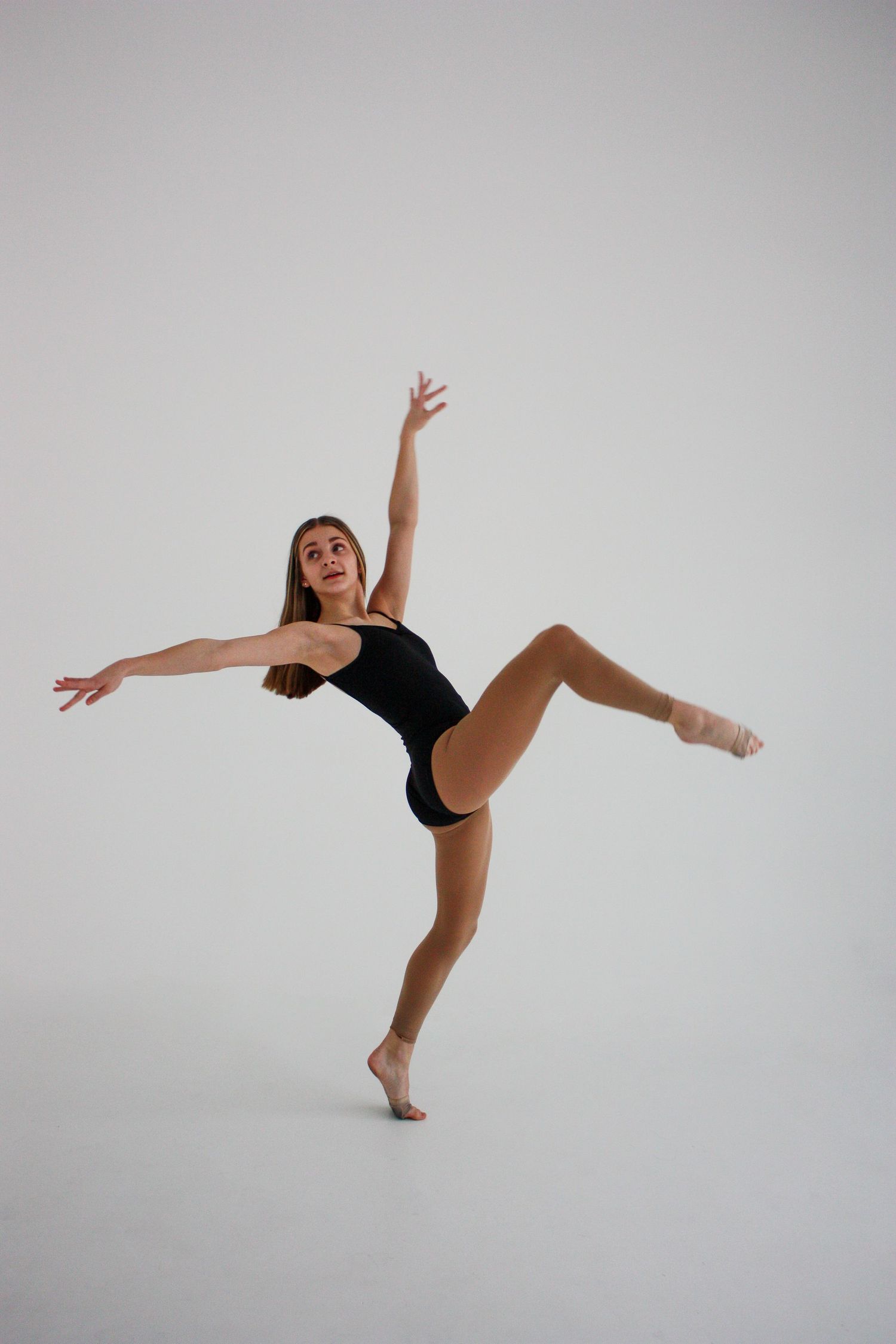 Female ballet dancer in black leotard balancing on one leg with arms extended in a bright studio.