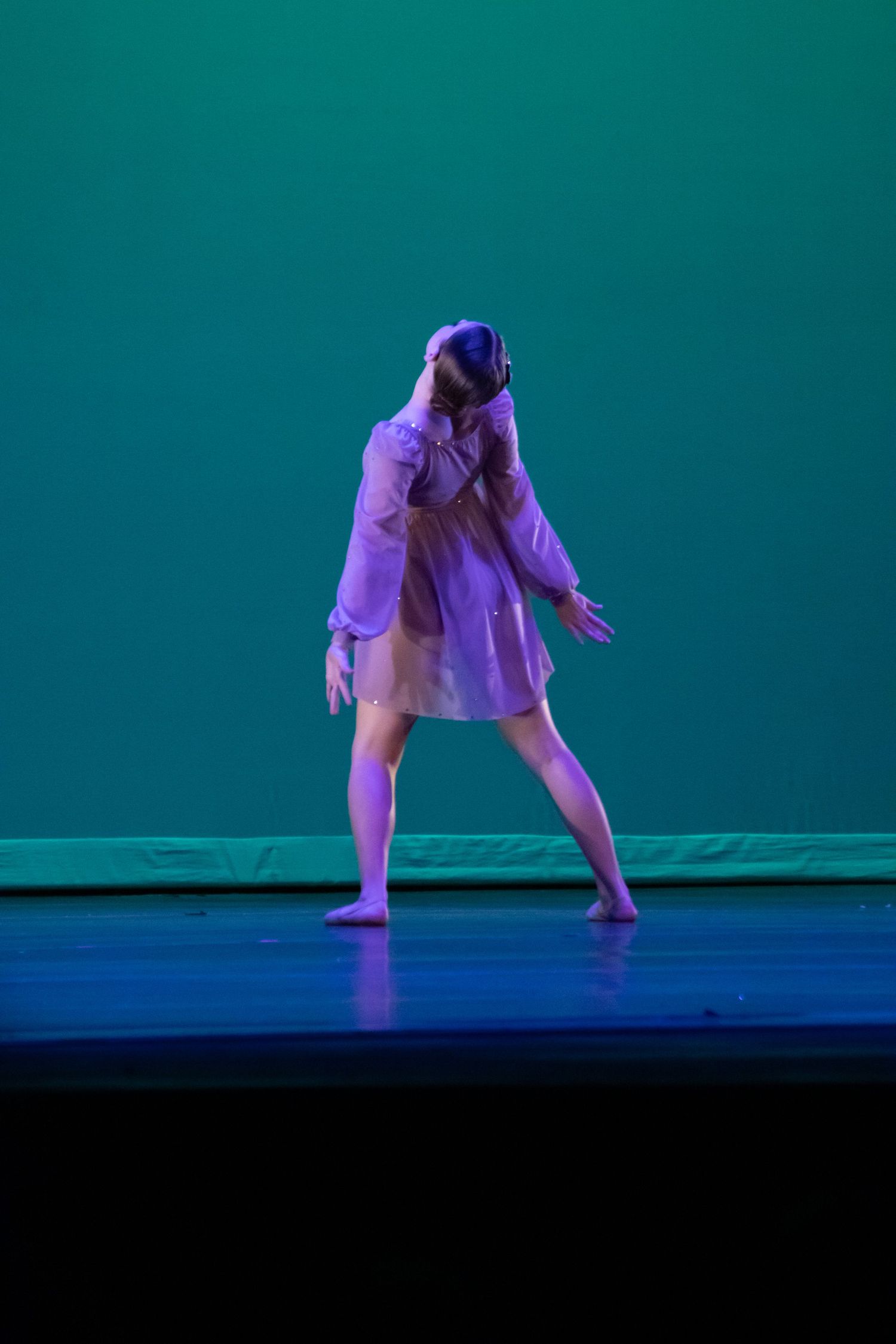 Solo dancer in white oversized shirt performs contemporary dance on a teal-lit stage.