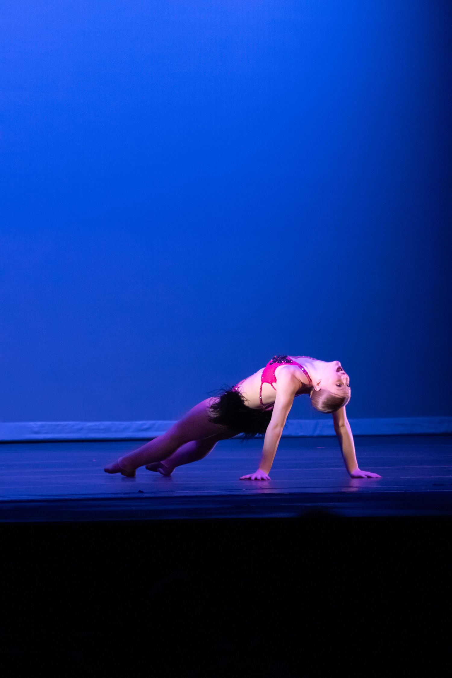 Young blonde dancer in pink and black costume performs low backbend on blue-lit stage.