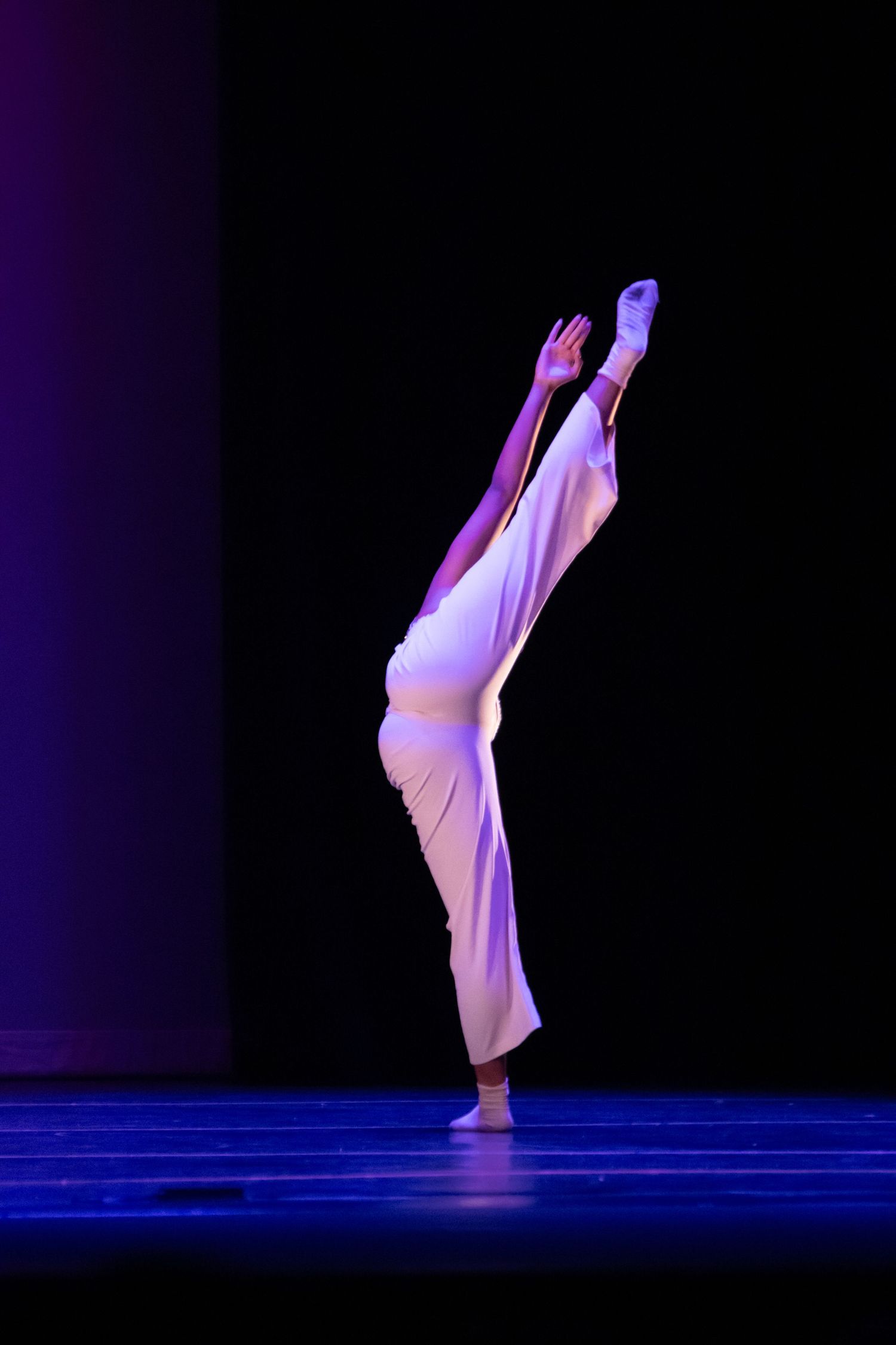 Dancer in flowing white outfit performs a high leg extension under purple stage lighting.