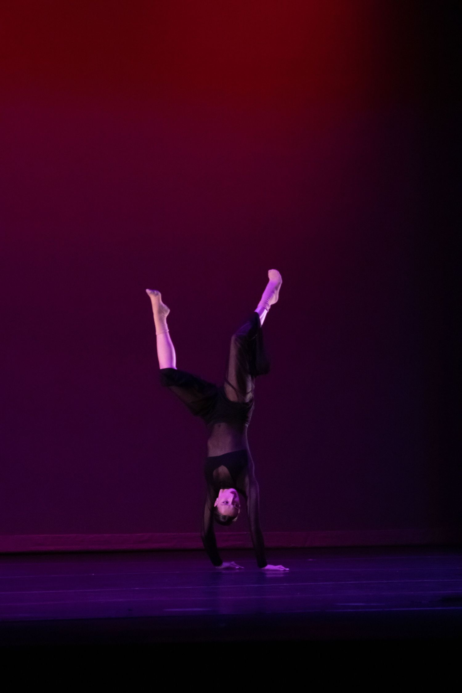 A dancer executes a perfect headstand with legs raised vertically under purple and red stage lights.