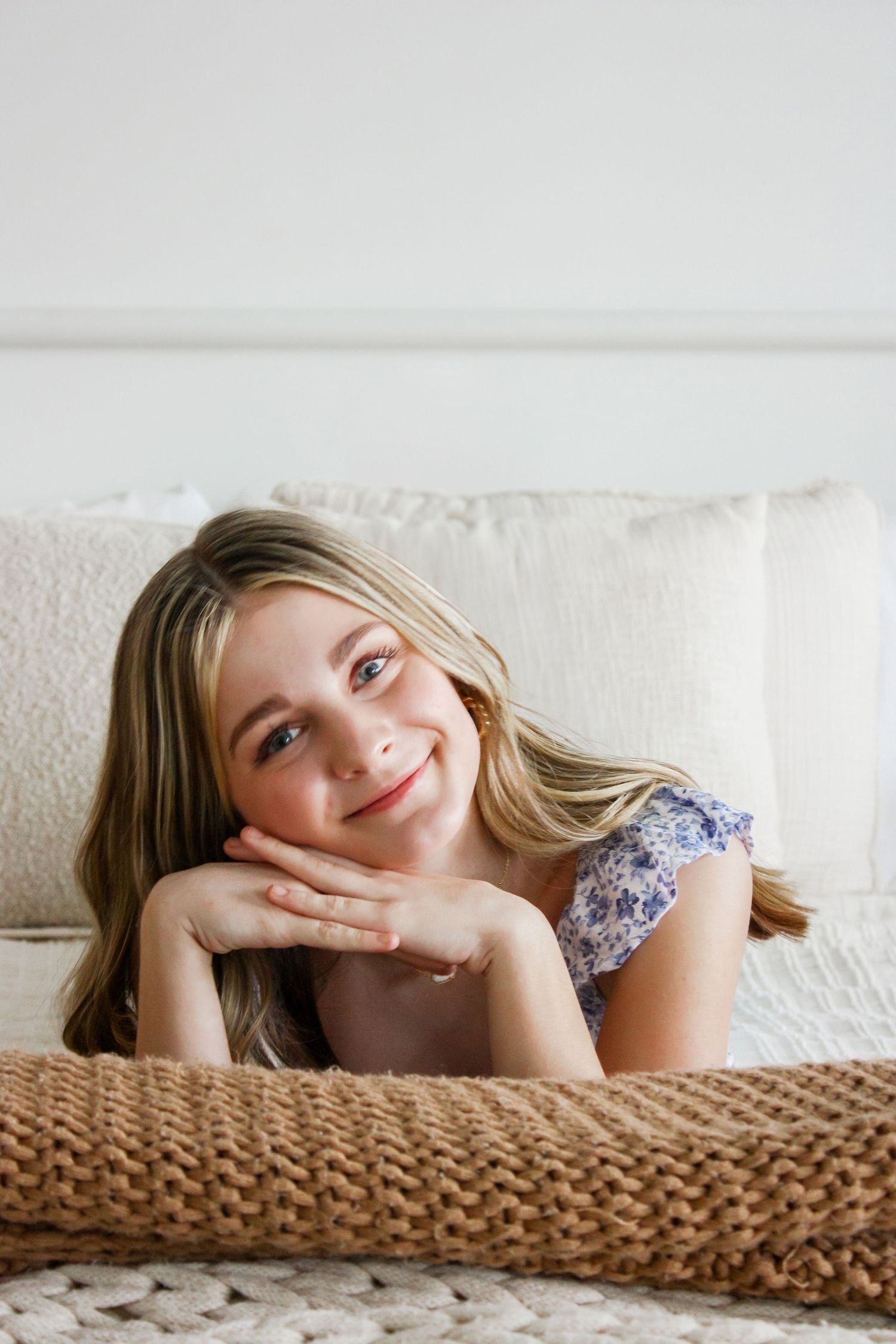Smiling young girl with long blonde hair resting chin on hands, lying on a cozy bed with white pillows.