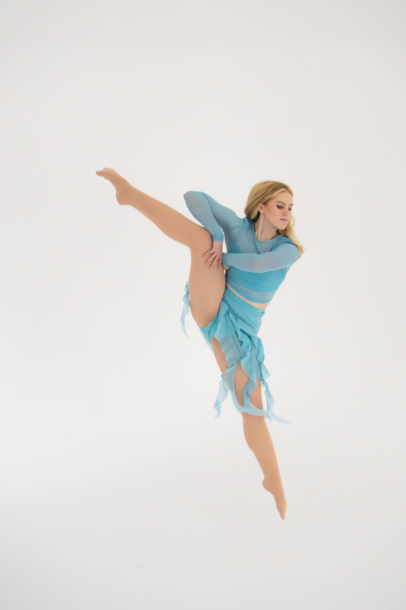 Blonde dancer jumping in turquoise costume with leg extended against white background.