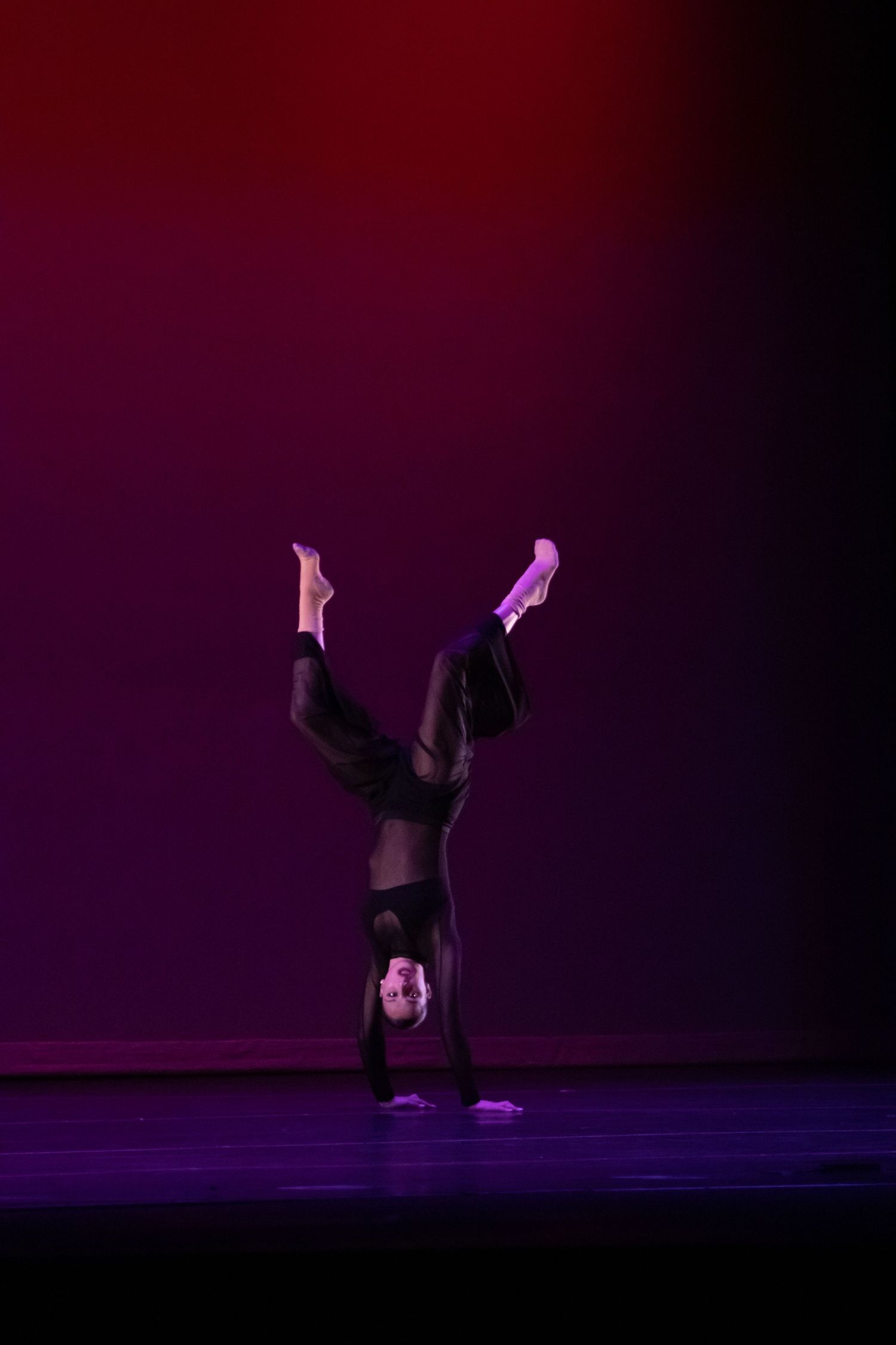 Dancer in black performing a headstand with legs split on a purple-lit stage.