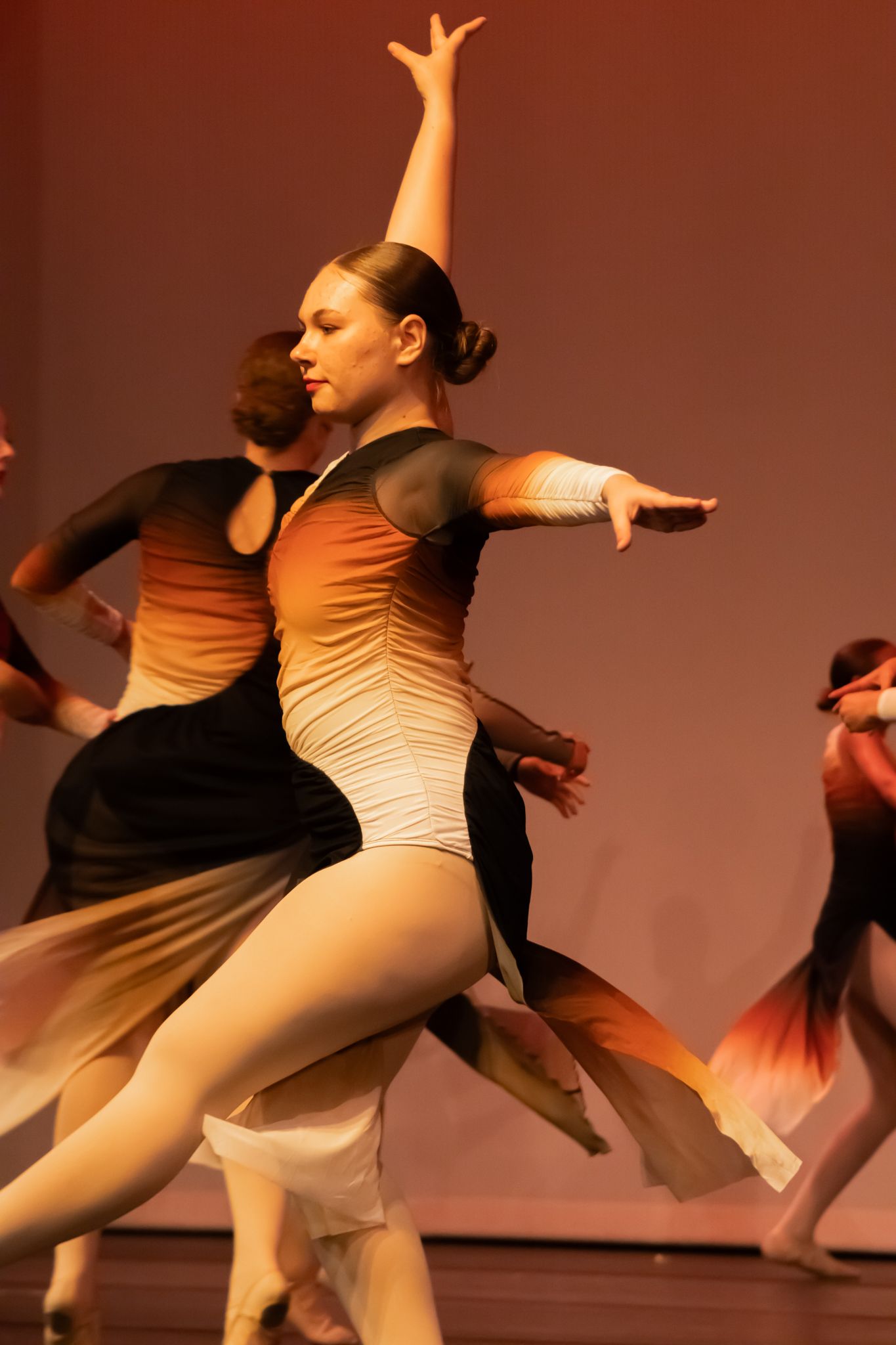 A female dancer in an orange and black gradient costume performs an expressive contemporary dance on stage.