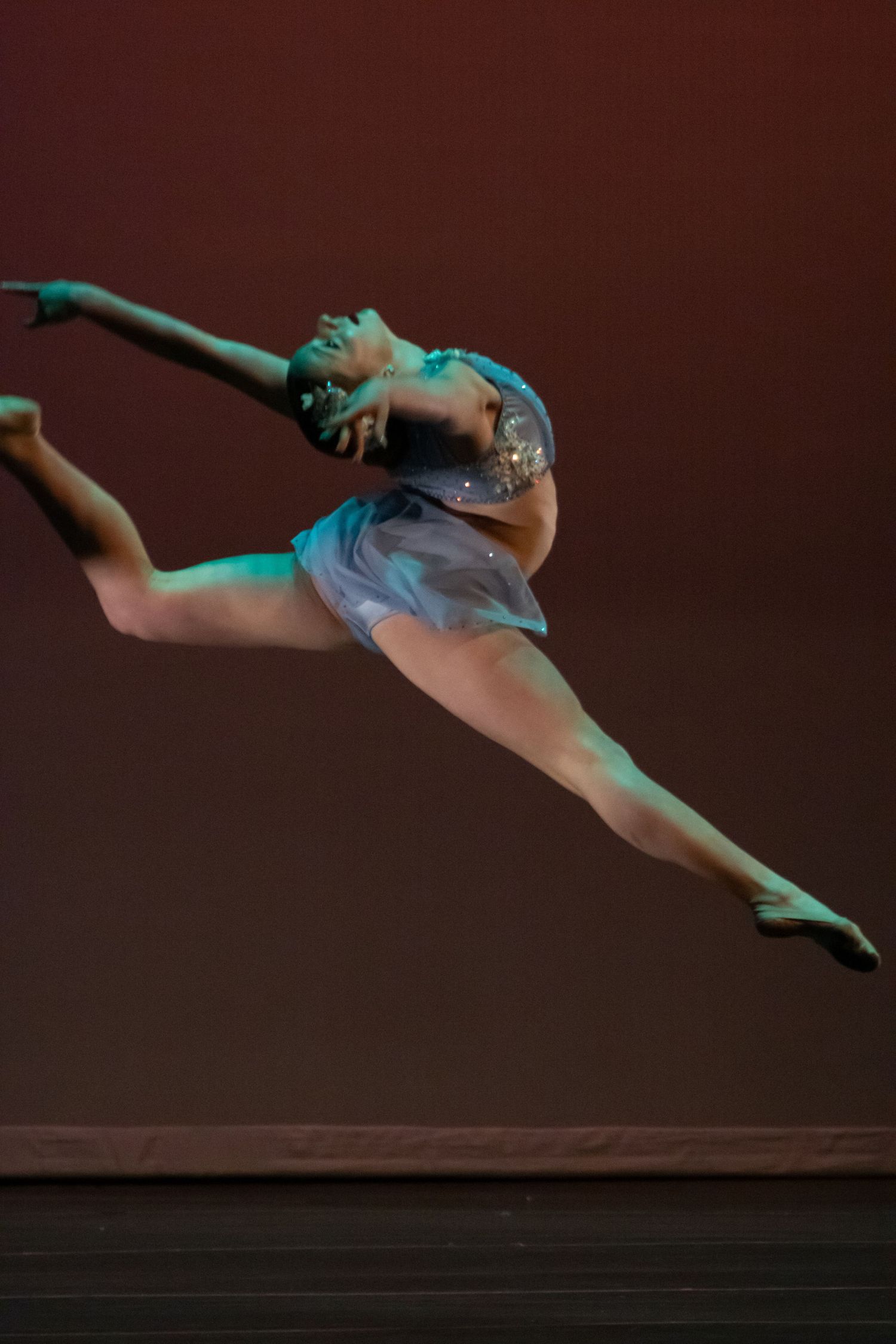 A dancer in a blue costume executes a stunning split leap on a warmly lit stage.
