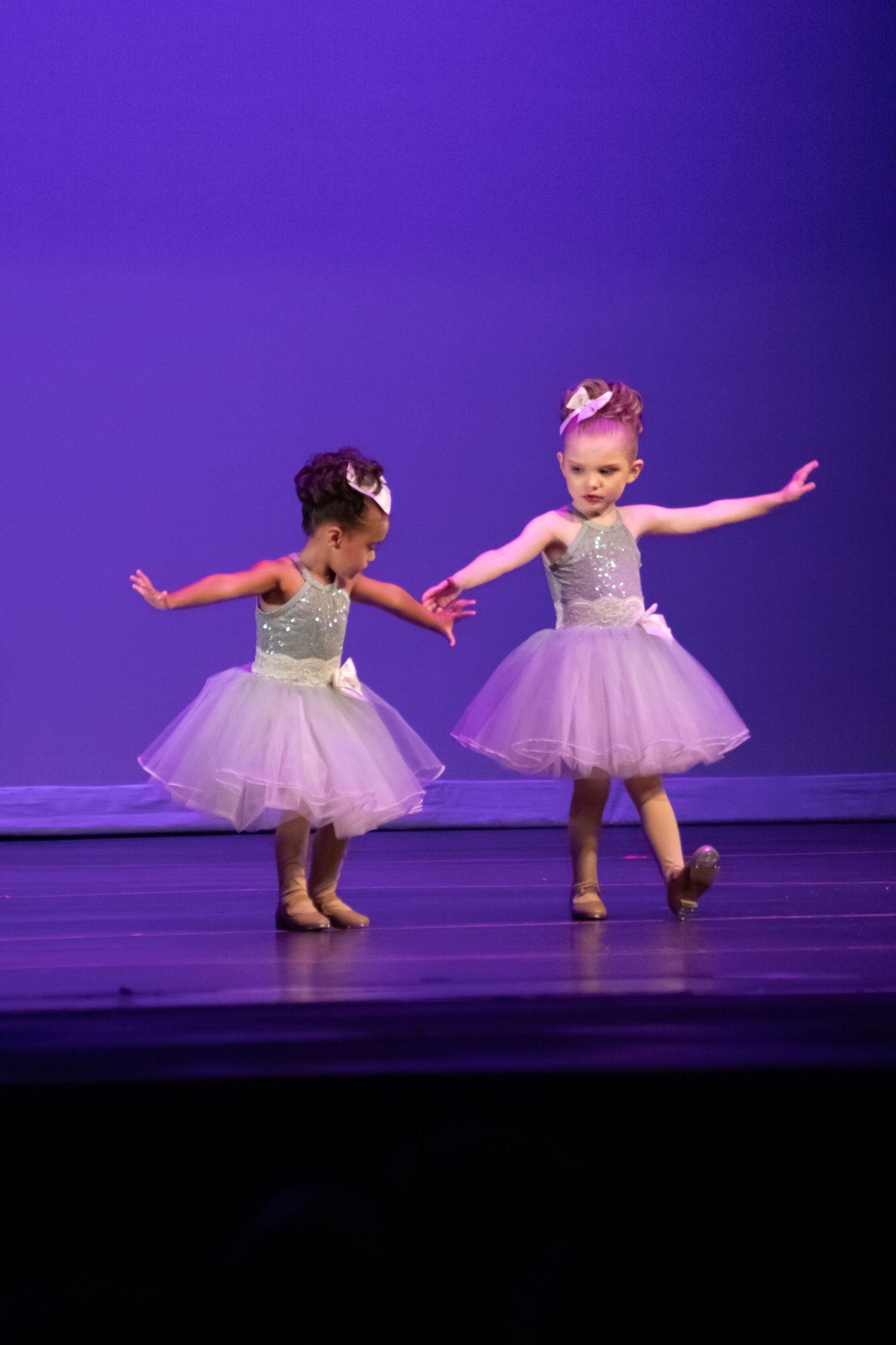 Two toddlers in silver and lavender tutus perform ballet on a purple-lit stage.