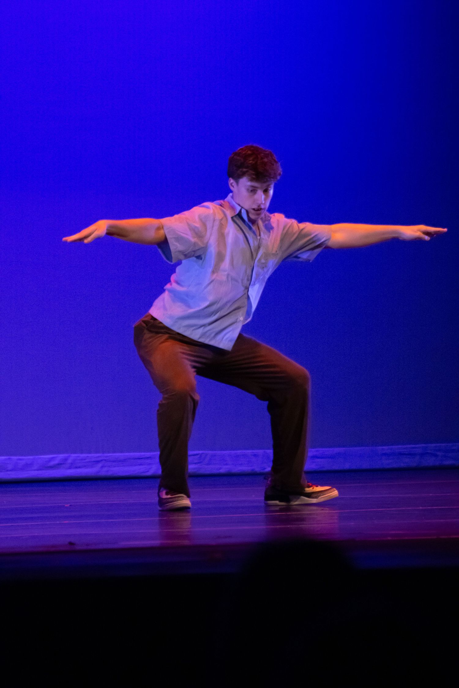 A young male dancer in casual clothes performs an energetic pose with arms outstretched on a blue-lit stage.