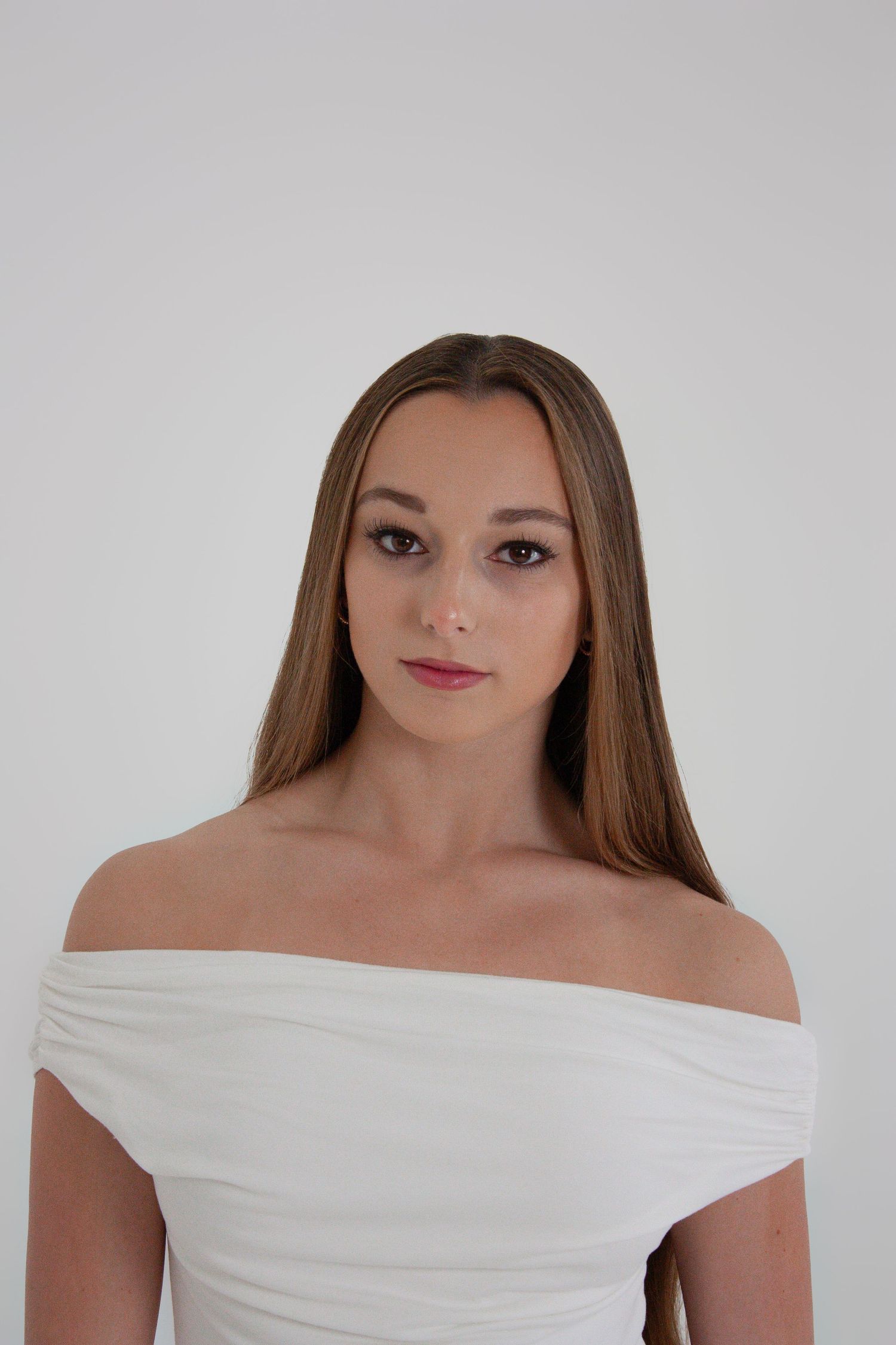 Young woman with long brown hair wearing an elegant white off-shoulder dress poses against a light gray background.