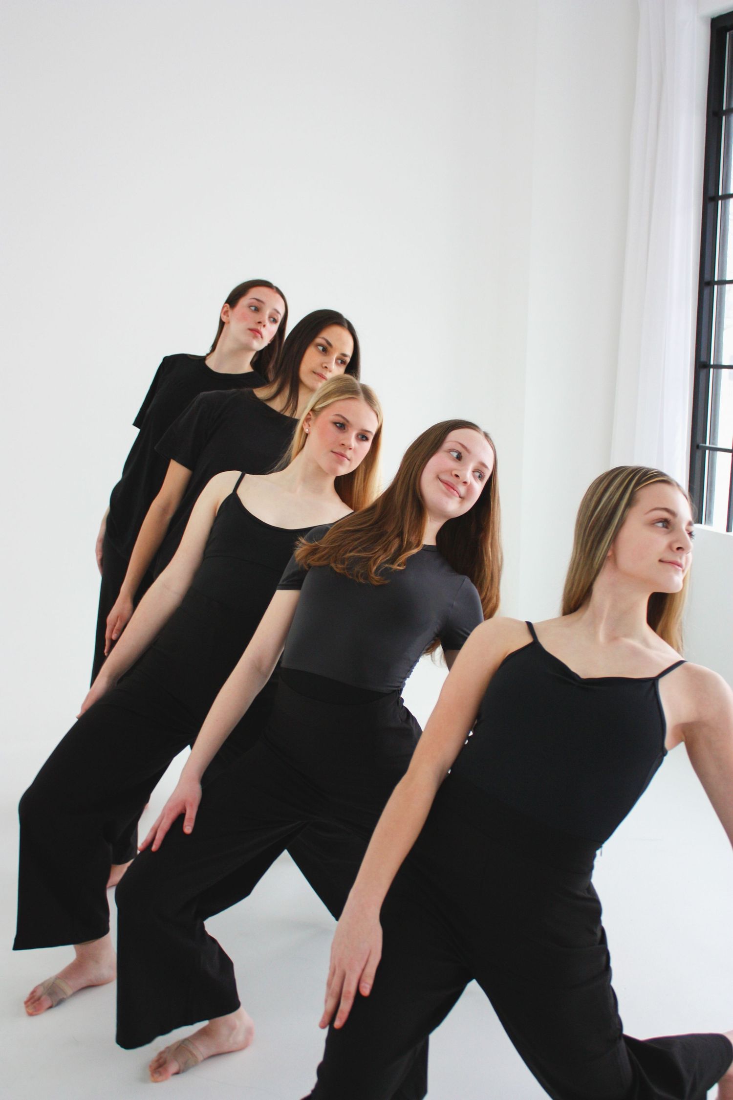 Five female dancers in all-black outfits pose in a diagonal formation in a bright white dance studio.