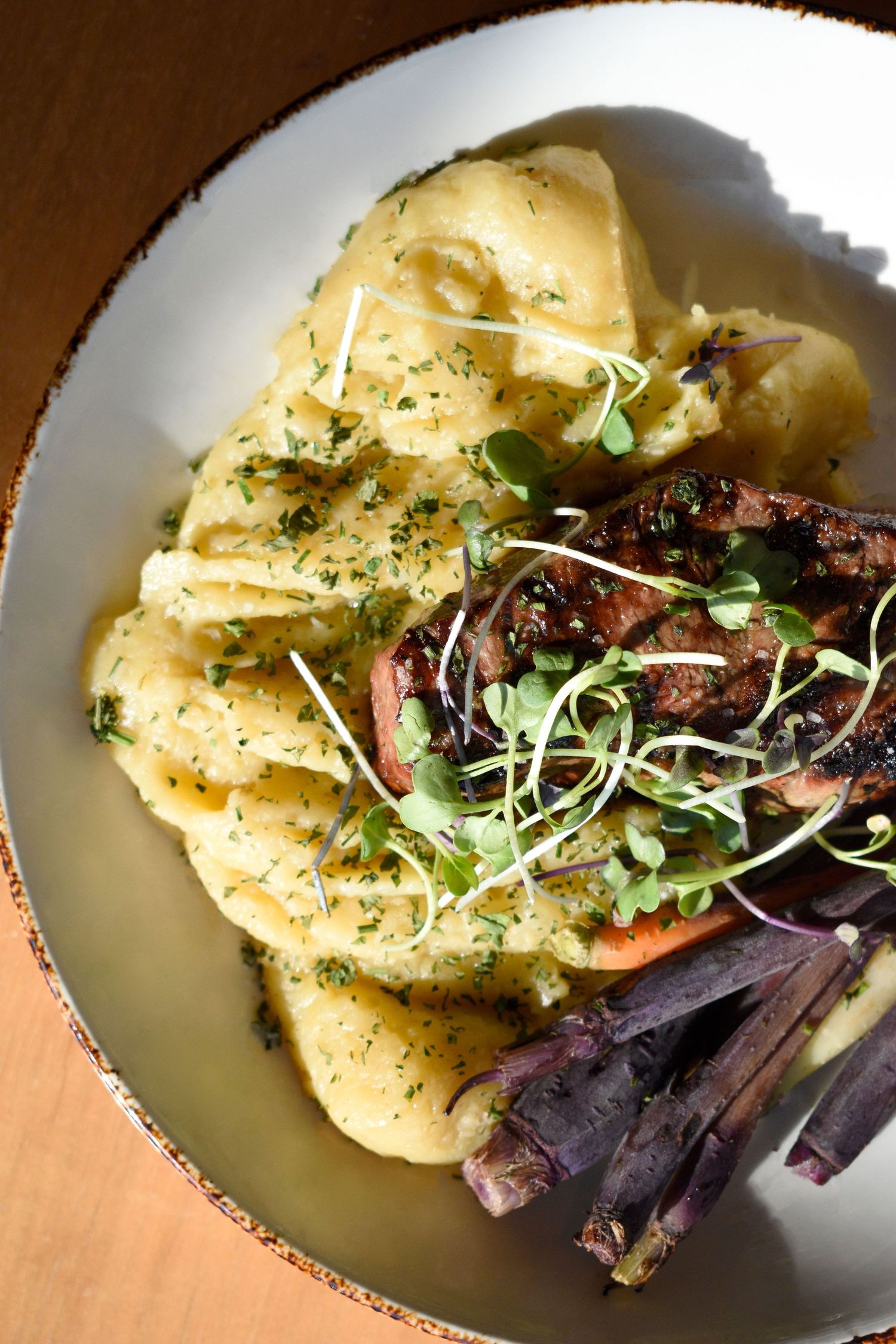 A plated dish of ravioli pasta topped with grilled steak, microgreens, and purple carrots.