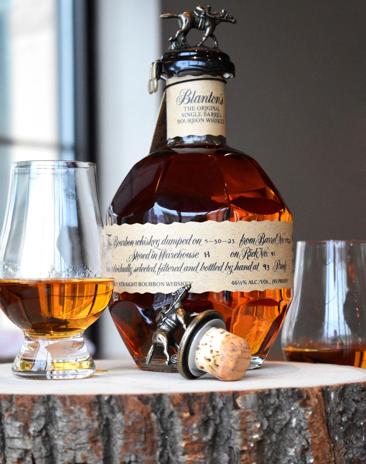 Blanton's Original Single Barrel Bourbon Whiskey bottle with Glencairn glasses and decorative stopper on a wood stump.