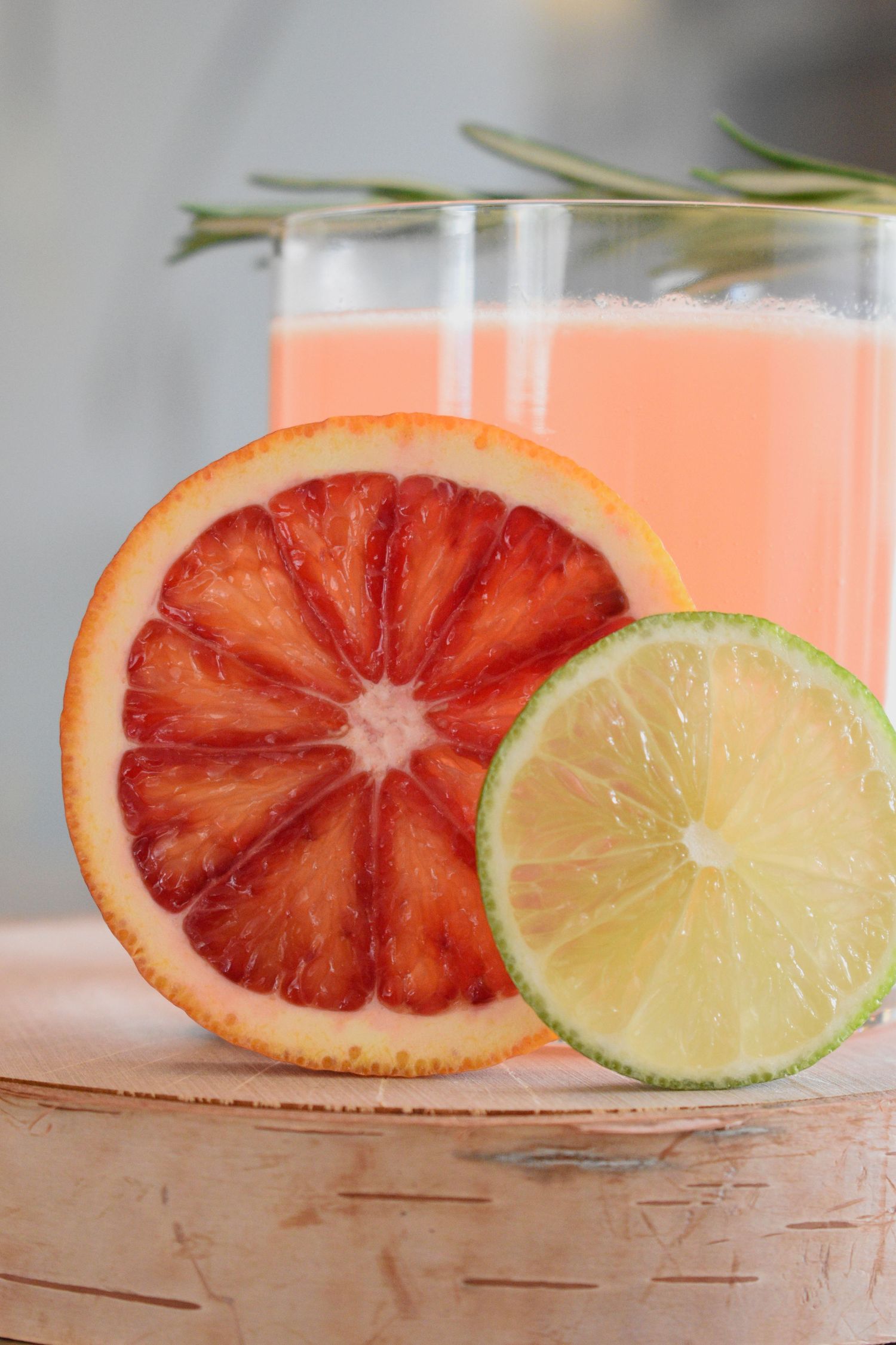 Sliced blood orange and lime half displayed in front of two pink grapefruit cocktails garnished with rosemary.