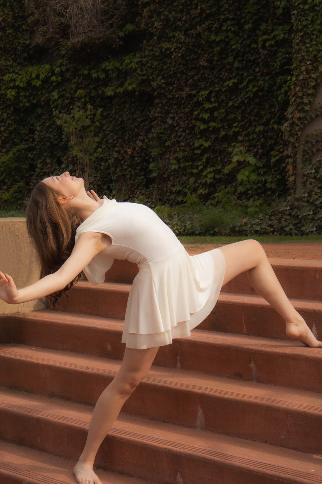 A barefoot dancer in a white dress performs a dramatic backbend on outdoor brick steps with ivy backdrop.