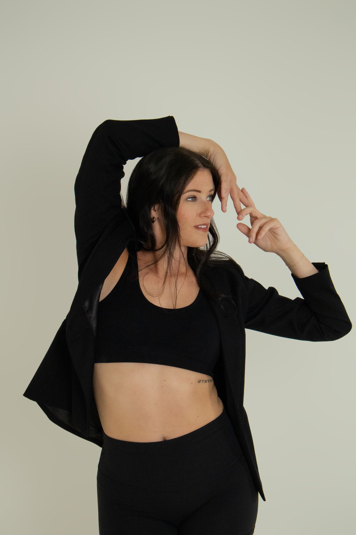 Woman in black crop top, blazer, and leggings posing in studio.