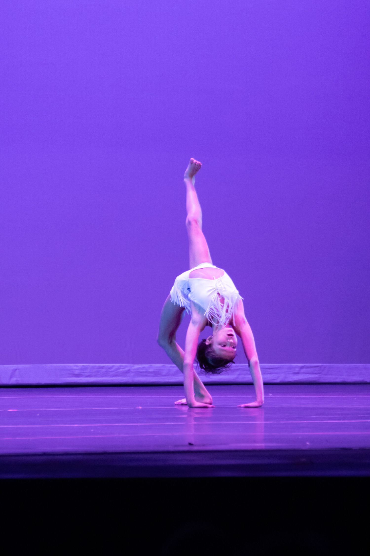 A young dancer in white performs a back walkover with one leg extended high on a purple-lit stage.