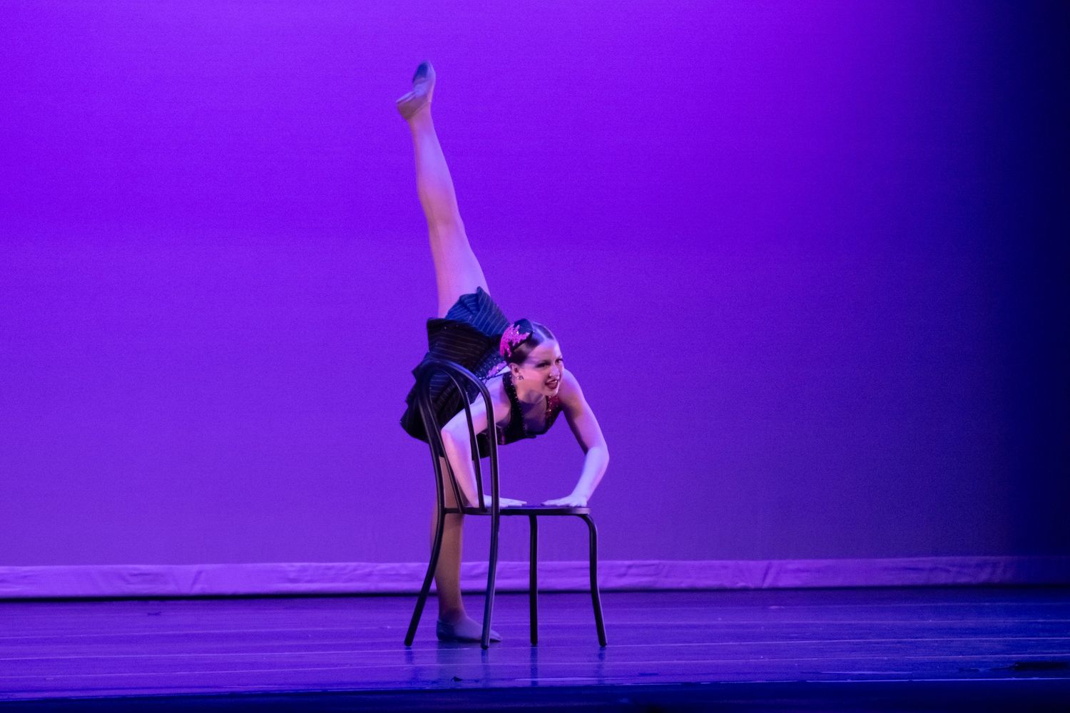 A flexible dancer performs an extreme backbend on a chair with one leg raised high on a purple-lit stage.