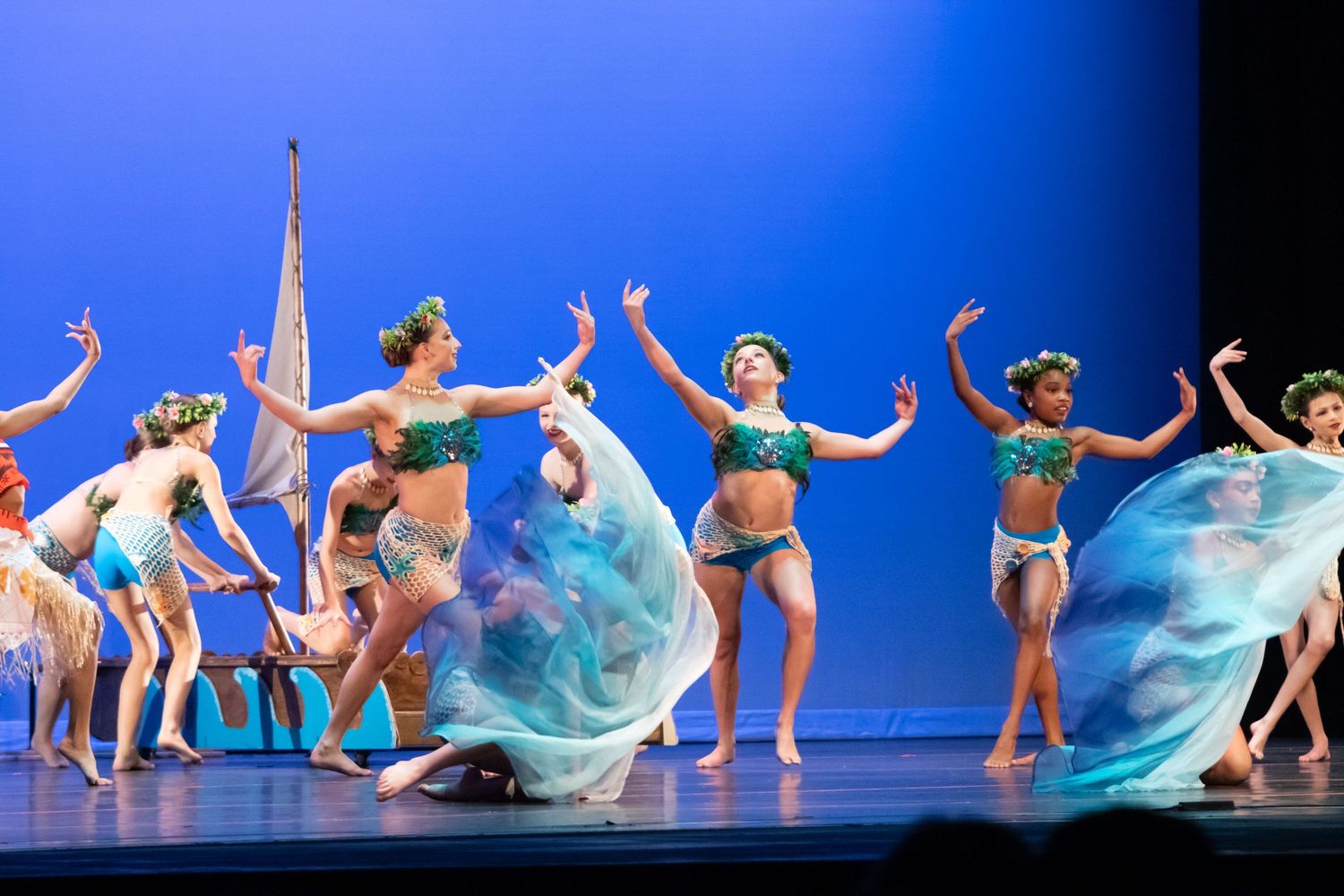 A group of dancers perform in ocean-themed costumes with flowing blue skirts on a bright stage.
