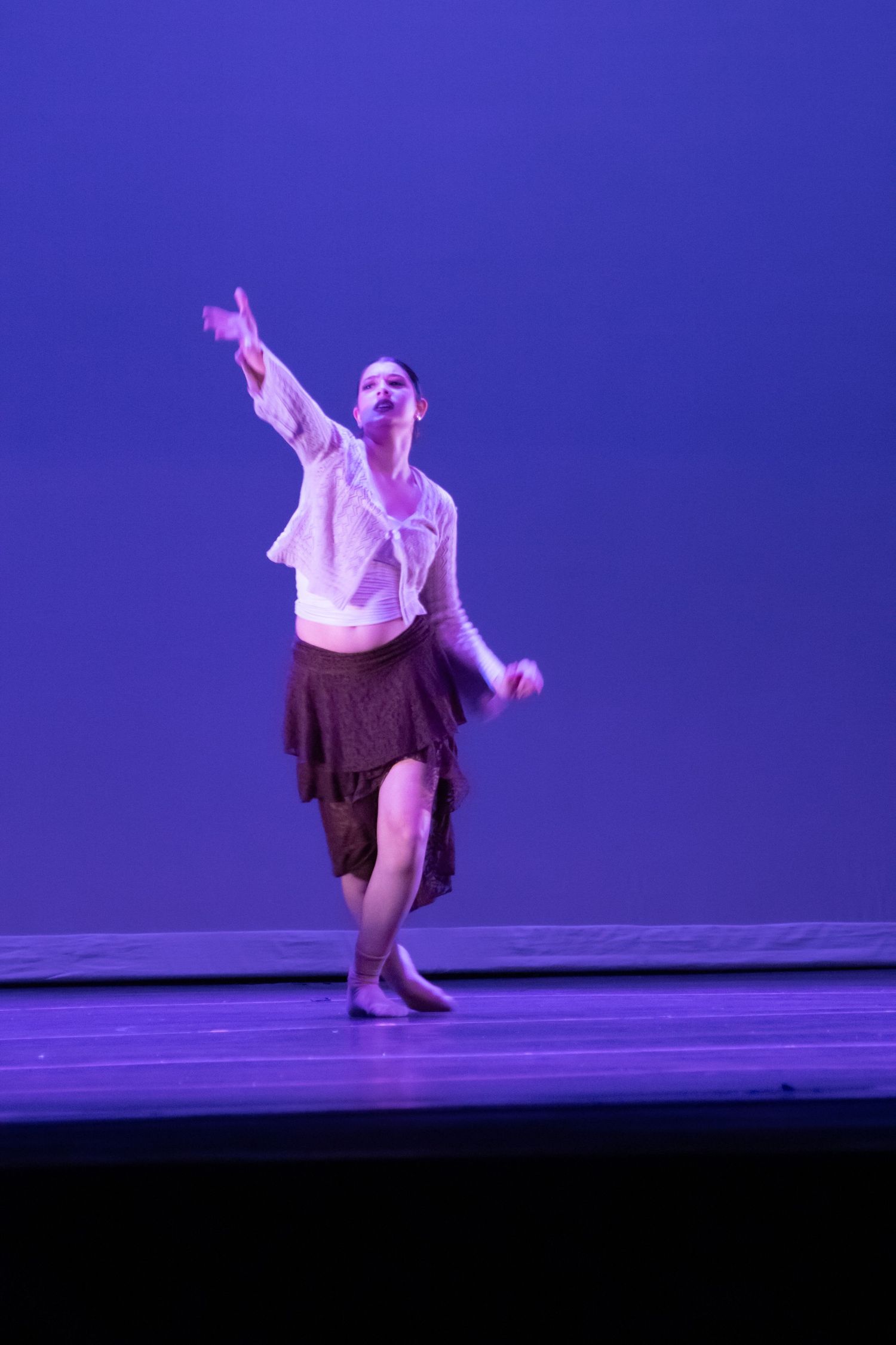 Female dancer in white blouse and brown skirt reaches arm upward on purple-lit stage.