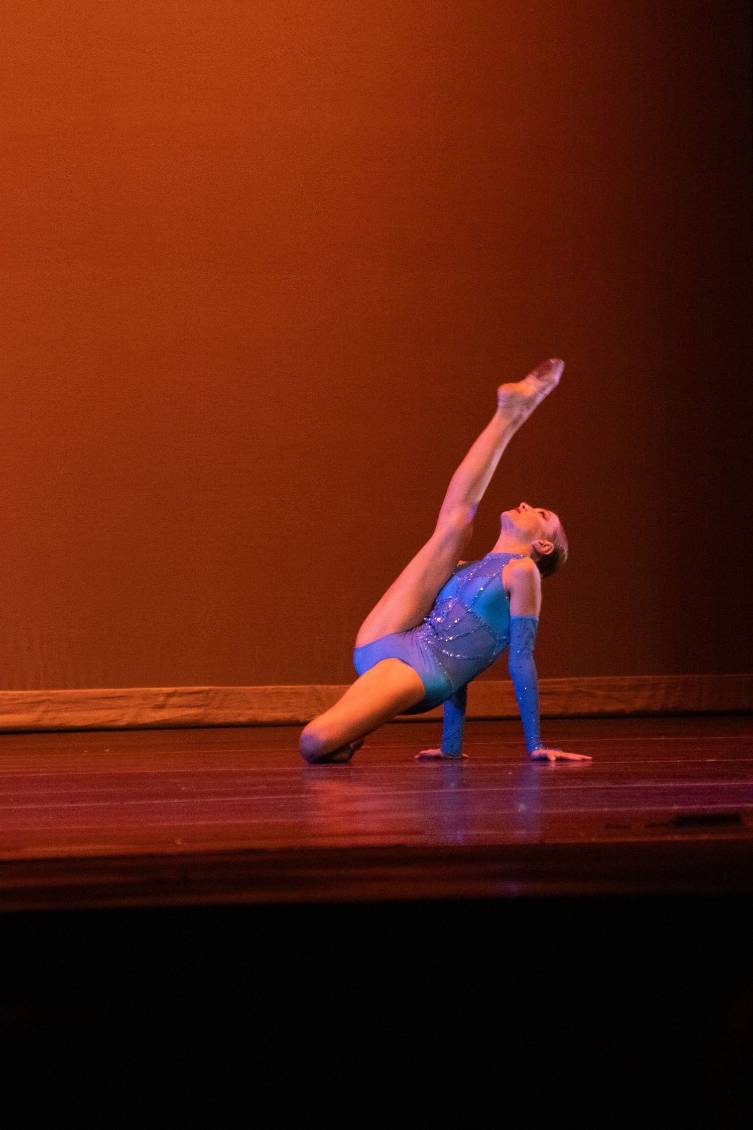 A young gymnast in a blue leotard performs a graceful leg extension pose on a warmly lit stage.