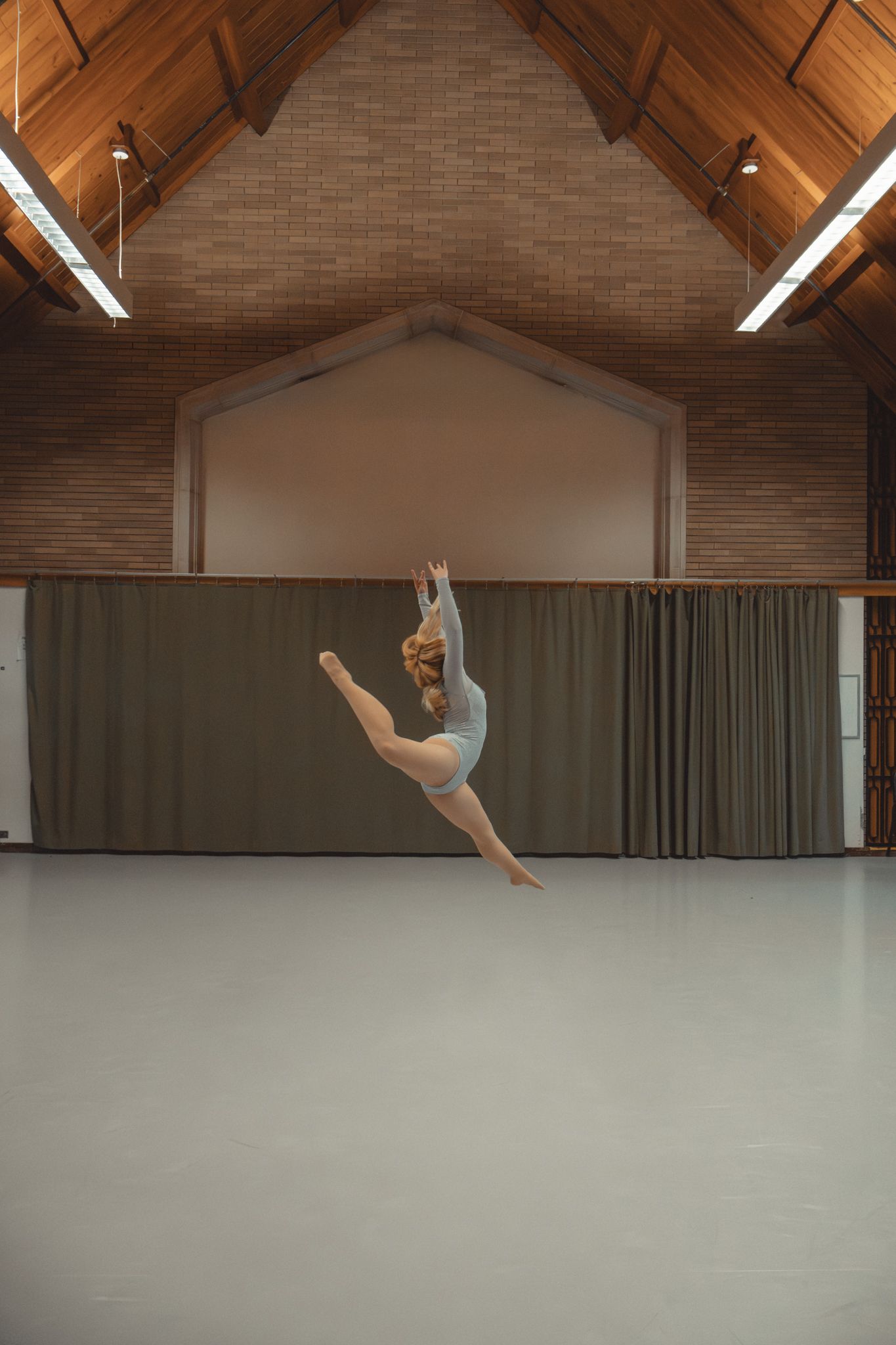 Blonde dancer in gray leotard performing a grand jete leap in a wooden dance studio.