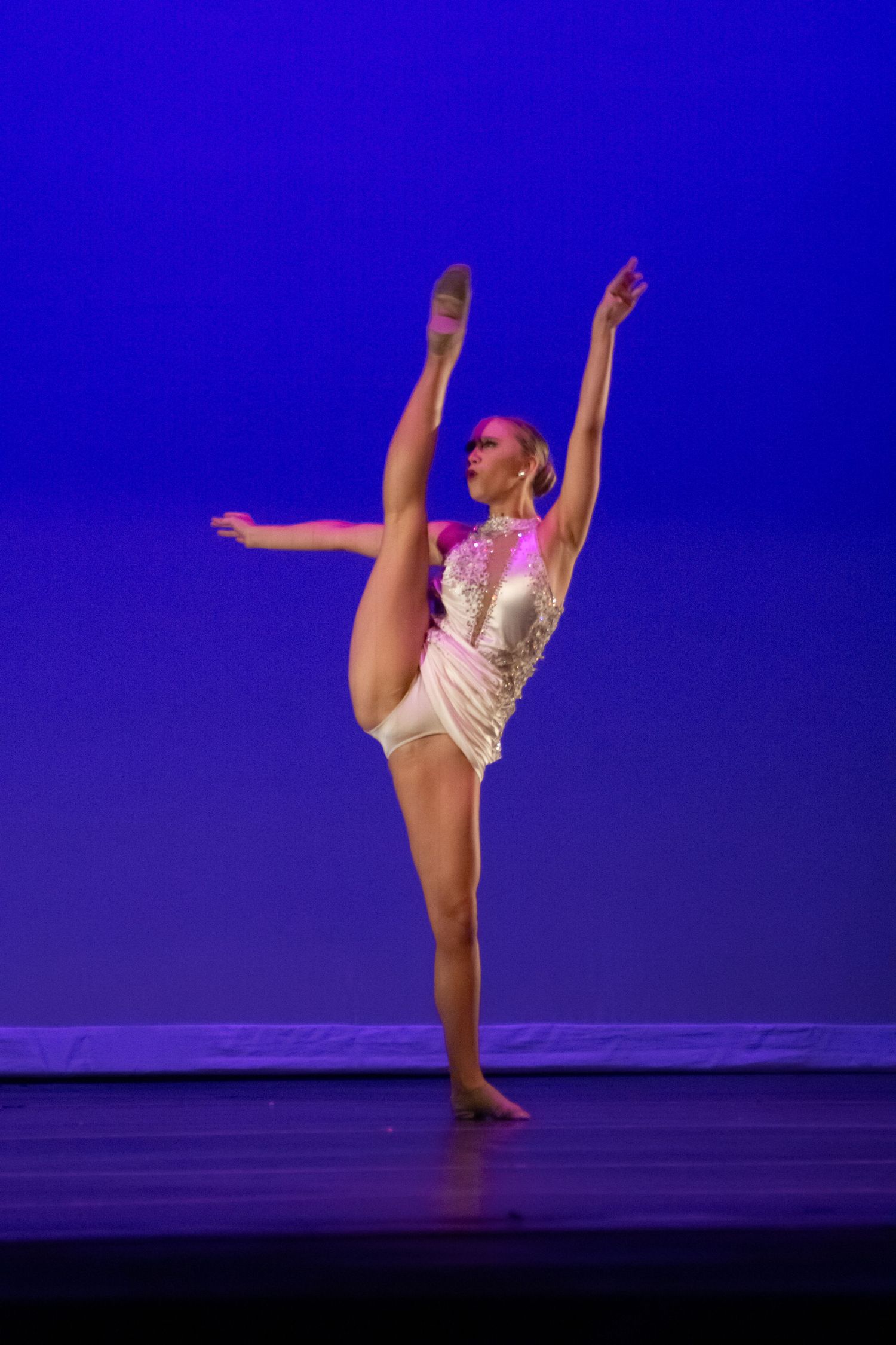 A female dancer performs an arabesque on stage in a white costume under blue stage lighting.
