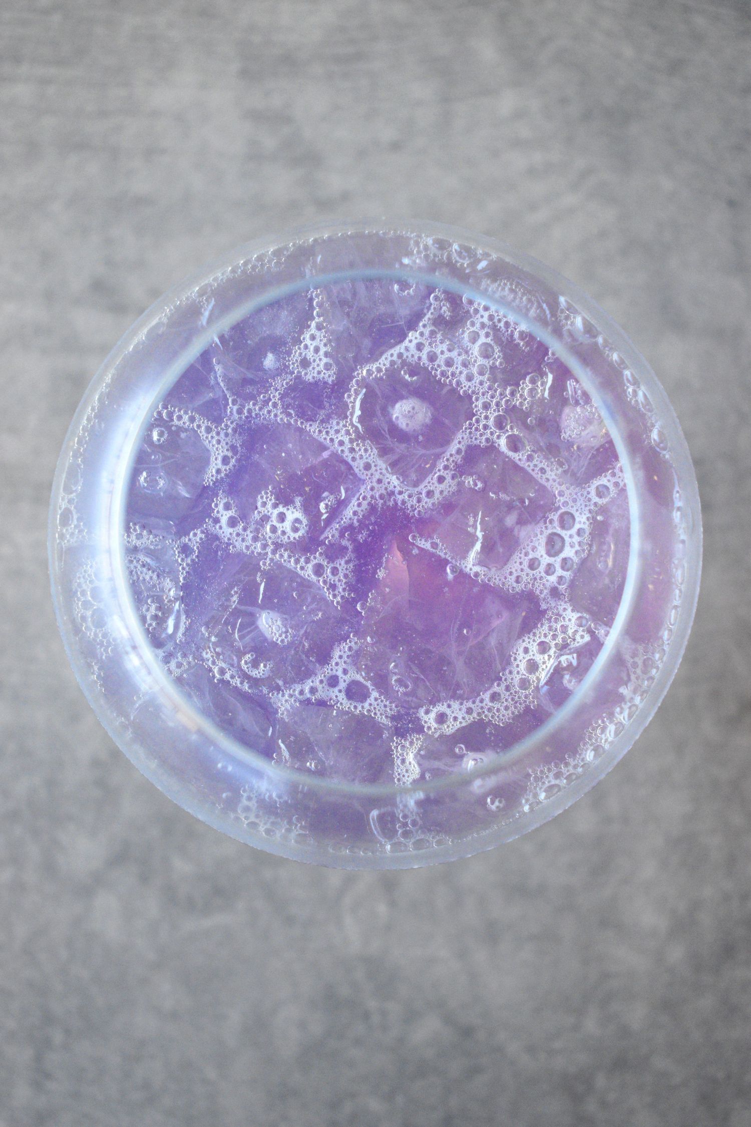 Overhead view of a purple-tinted cocktail with ice cubes and effervescent bubbles in a clear glass.