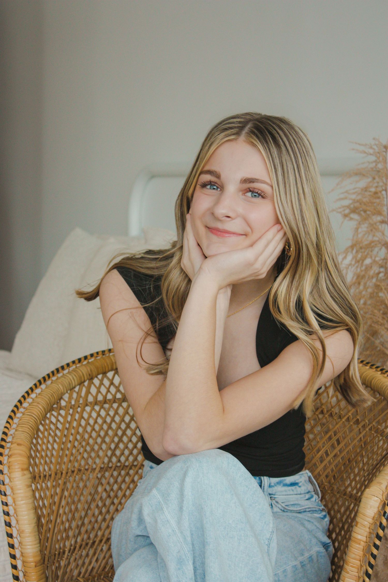 Young blonde woman smiling in a wicker chair, wearing a black top and light jeans in a cozy neutral-toned room.