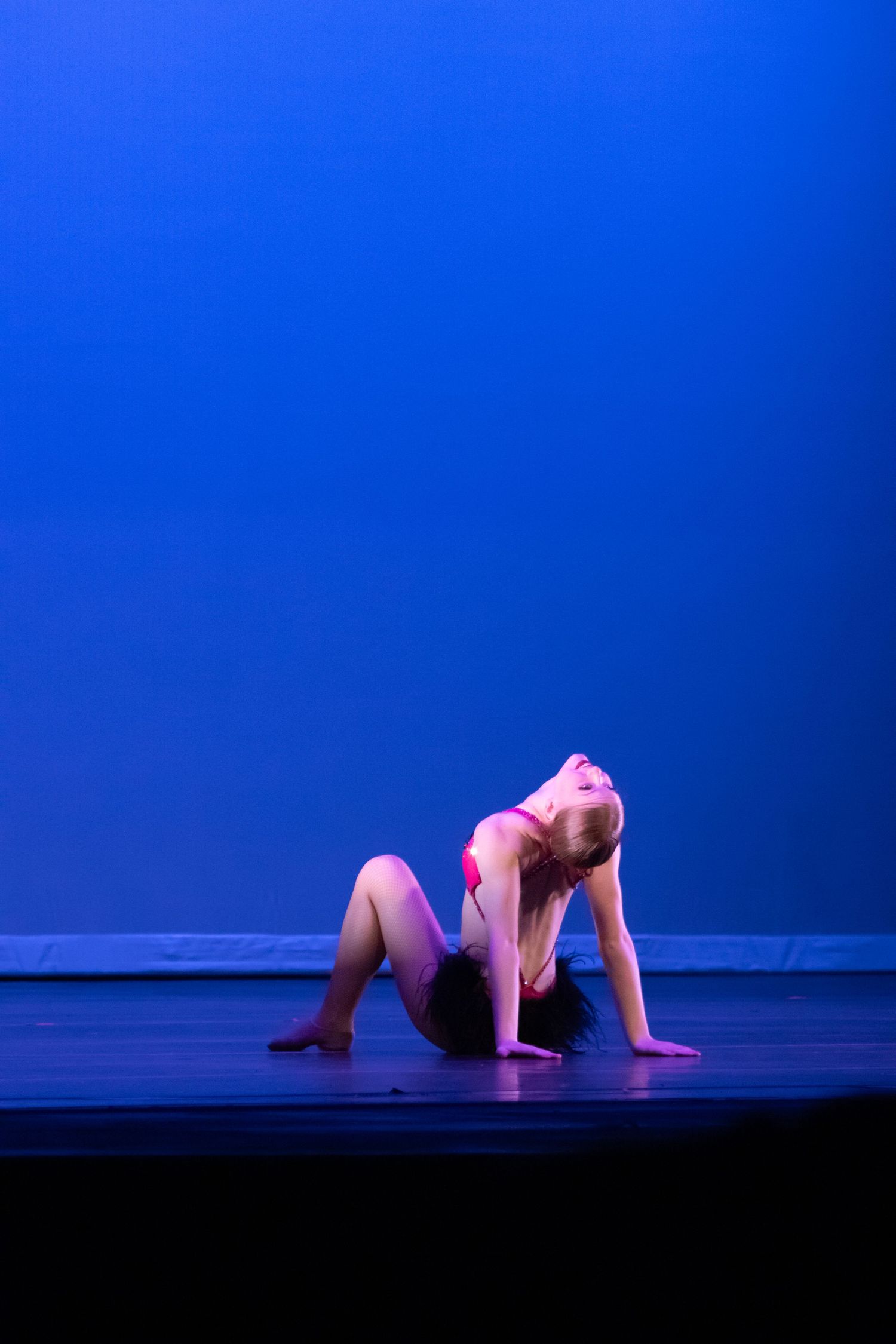 Young blonde dancer in pink costume performs floor backbend under blue stage lighting.