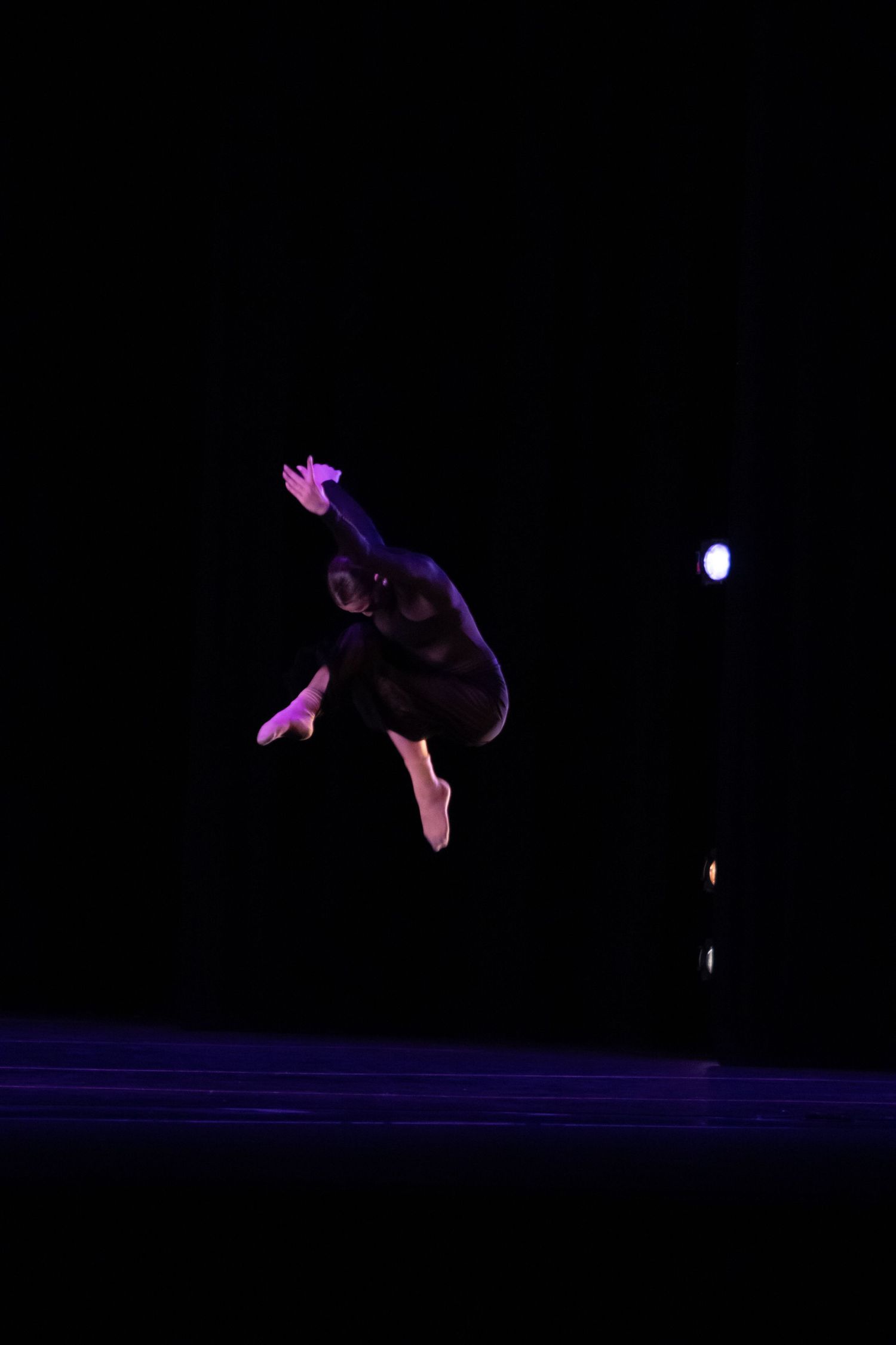 A dancer leaps dramatically against a dark stage backdrop with purple lighting.