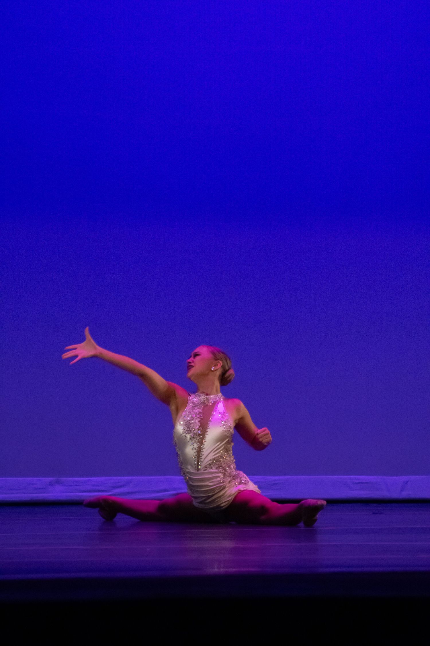 A dancer in a sequined costume performs splits on a blue-lit stage.