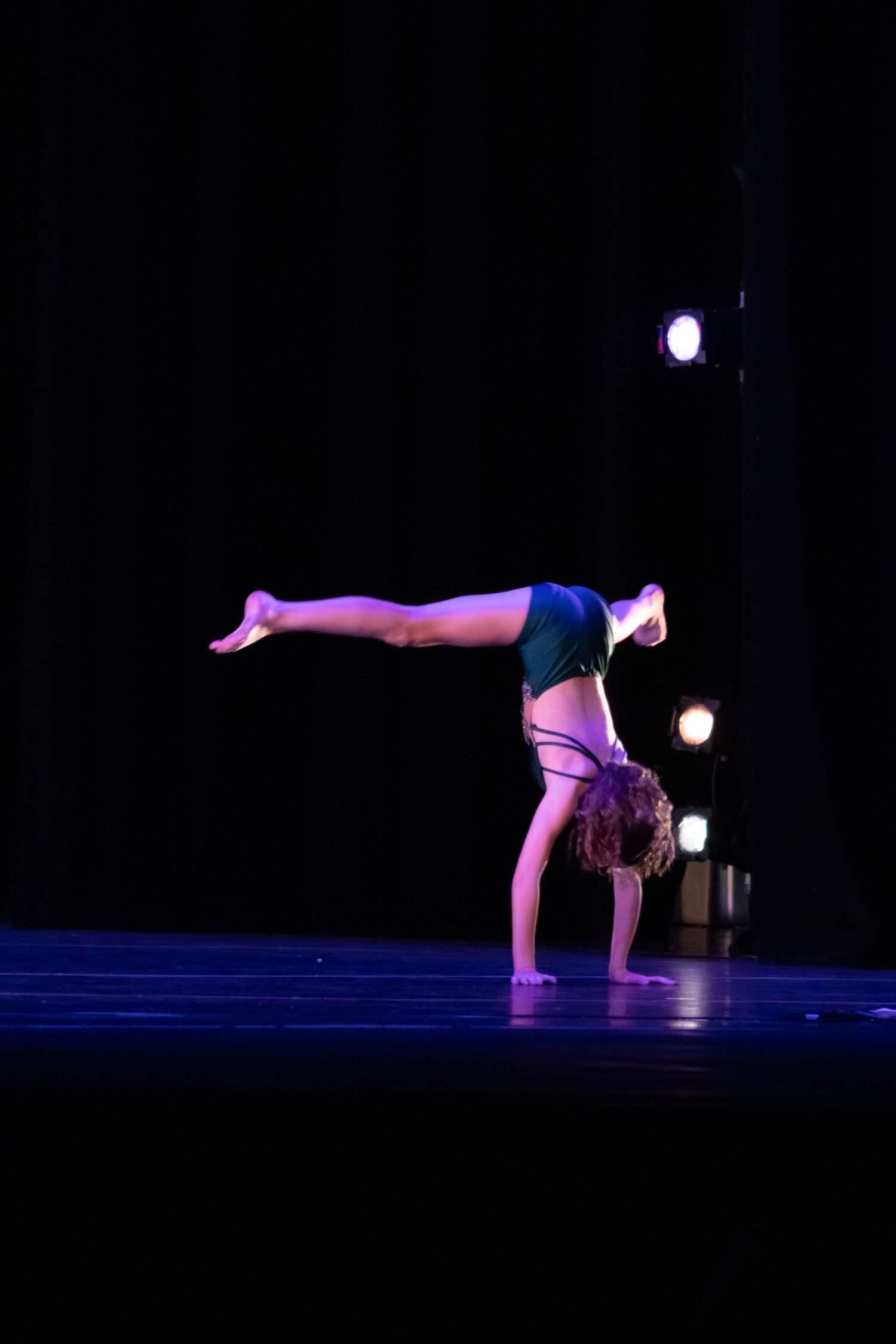 A flexible dancer performs a handstand with legs split on a dark stage lit by spotlights.