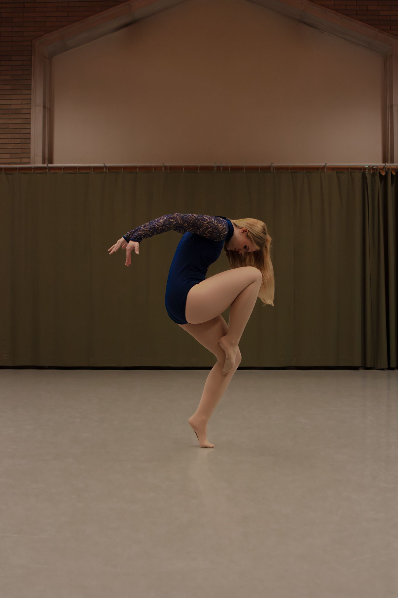 Blonde dancer performing dynamic leap in blue leotard in studio dance hall.