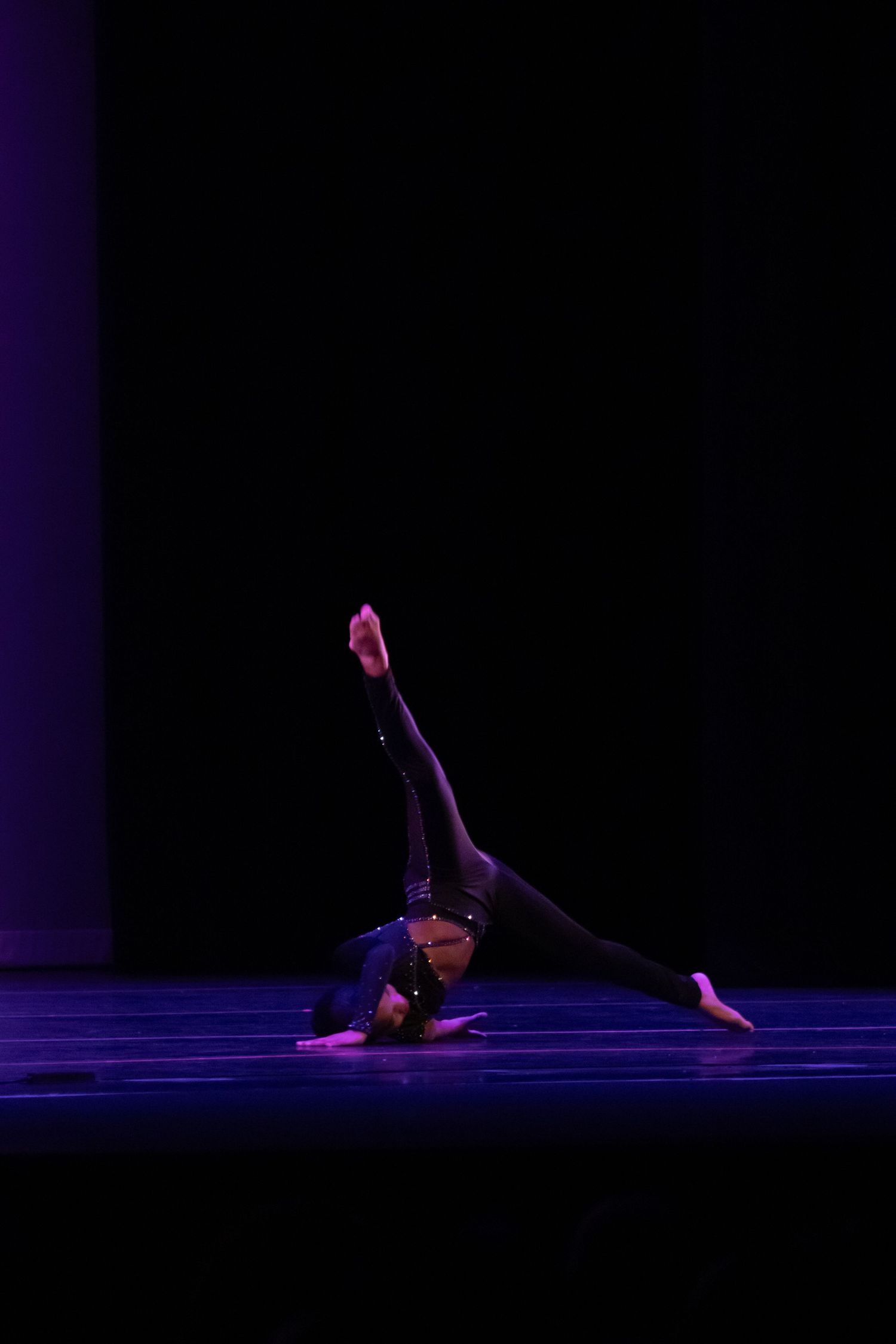 Female dancer in all-black performs an acrobatic floor pose on a dimly lit stage.