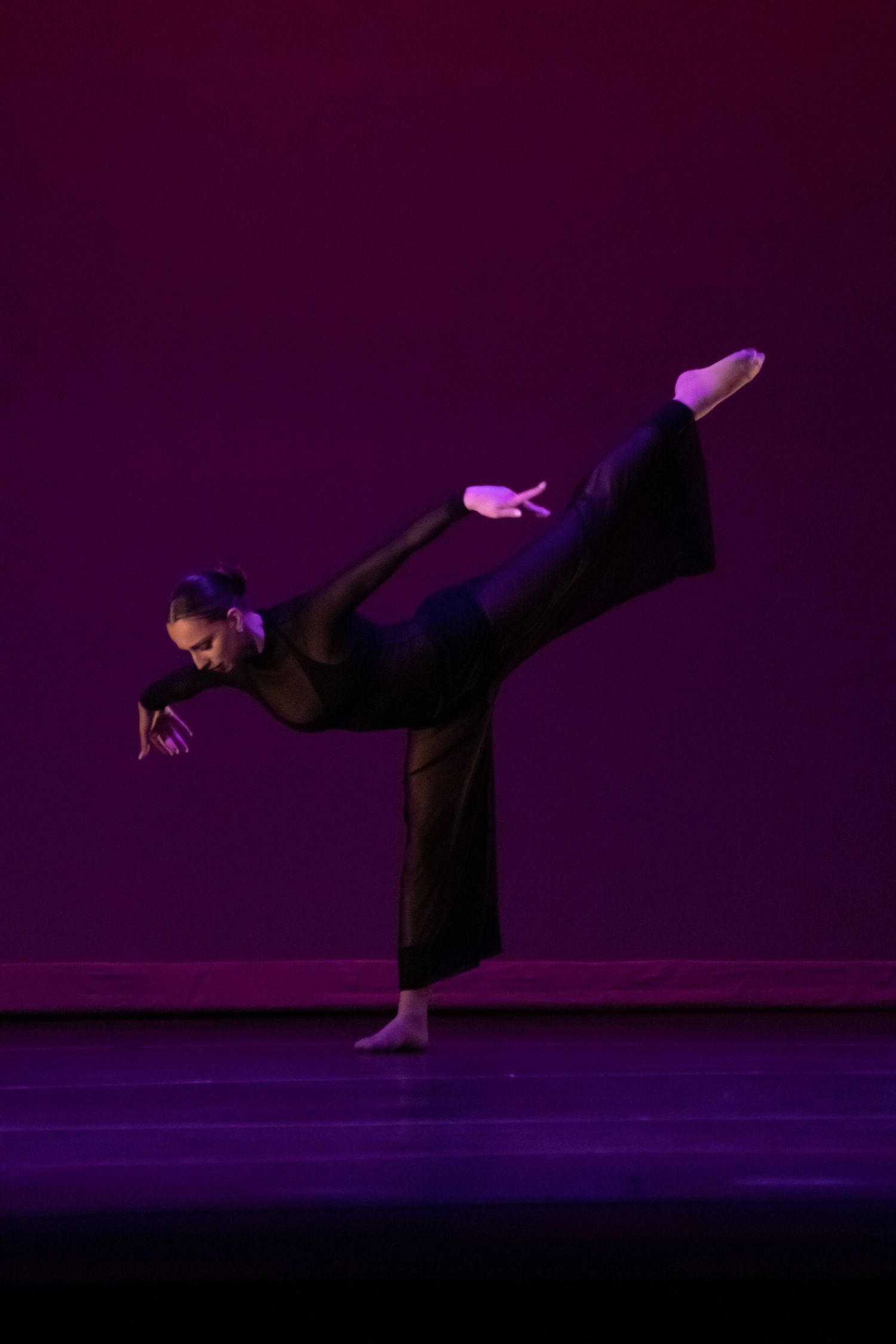 Dancer in black jumpsuit performs a high arabesque balance on a purple-lit stage.