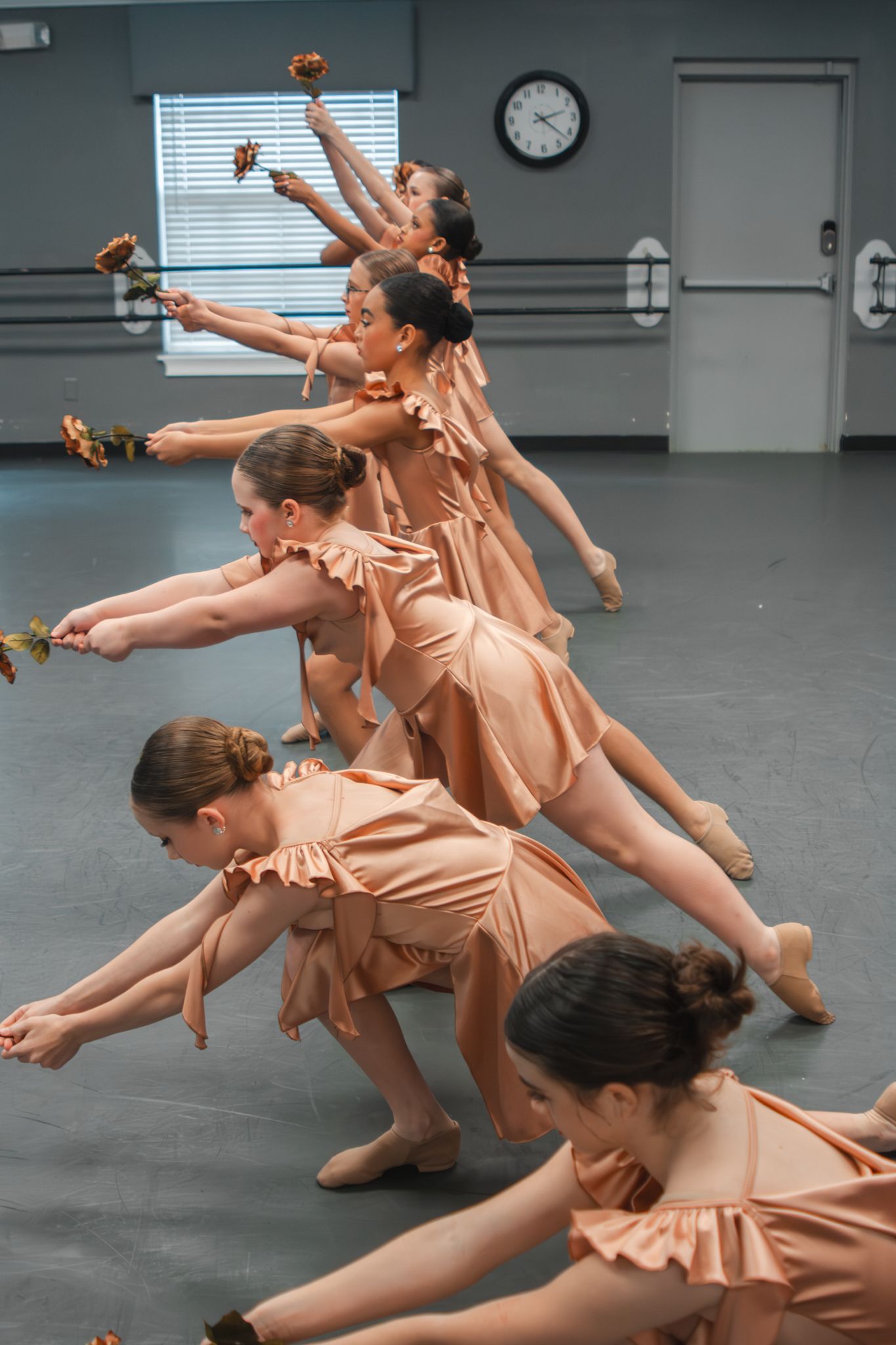 Young ballet dancers in peach costumes perform synchronized routine holding flowers in a dance studio.