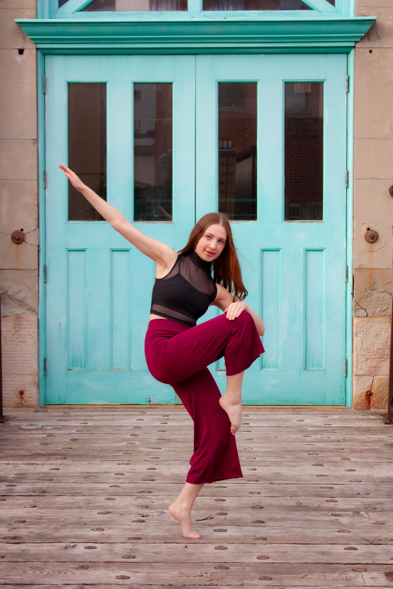 A dancer in burgundy pants and black top balances on one leg in front of a teal vintage door.