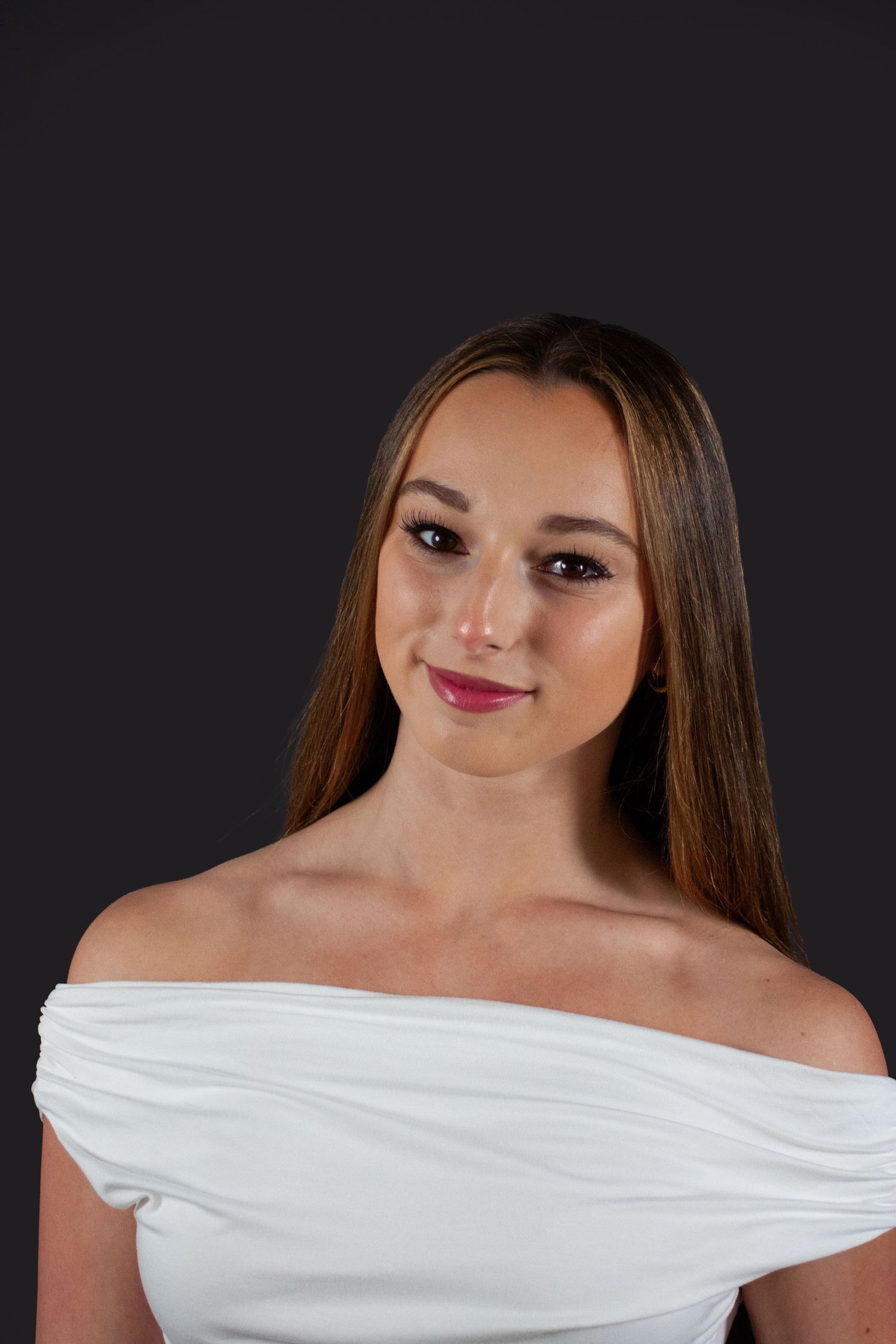 Young woman in white off-shoulder dress with red lips smiles in dark studio portrait photo.