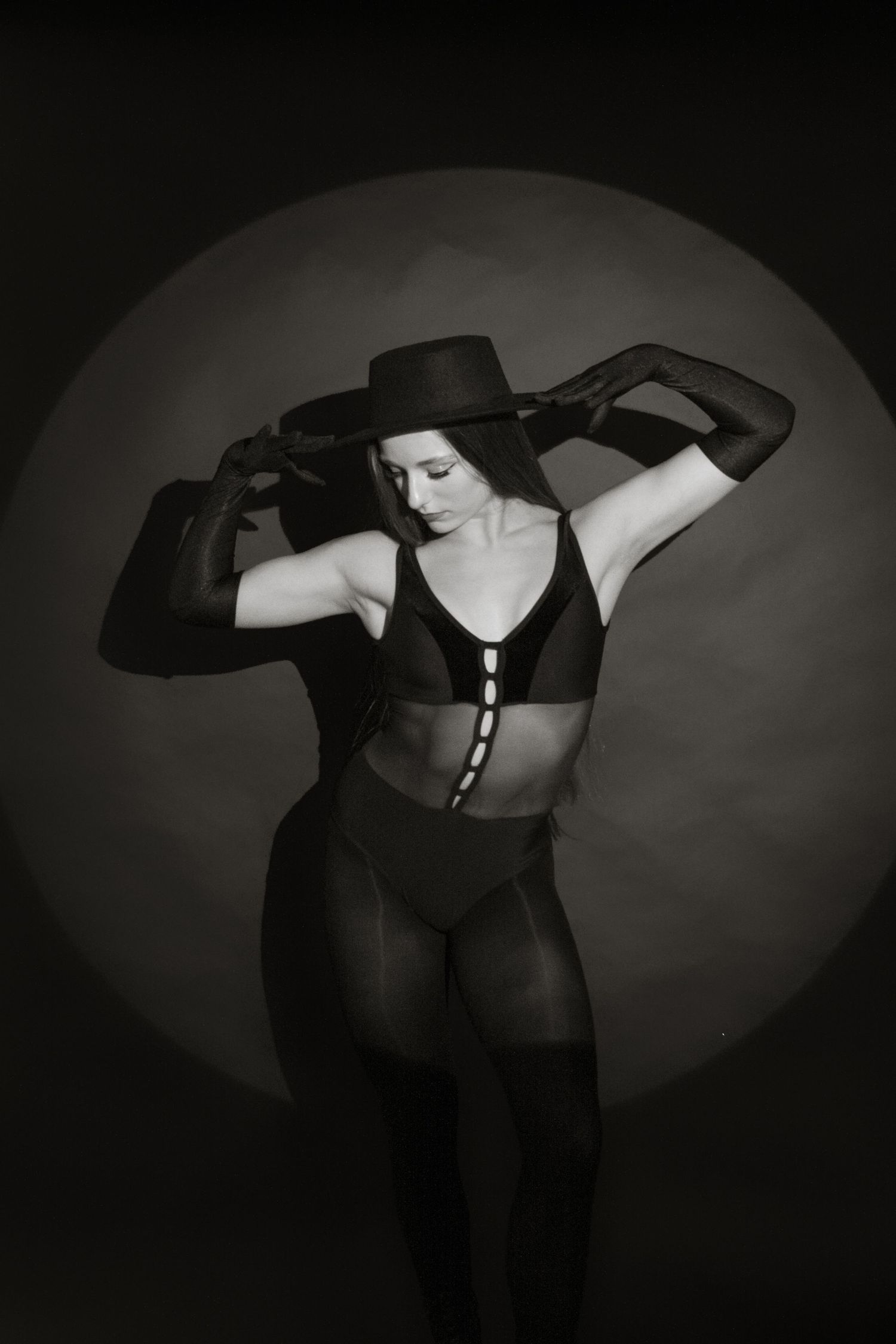 Black and white photo of dancer in black bodysuit adjusting wide-brim hat under circular spotlight.