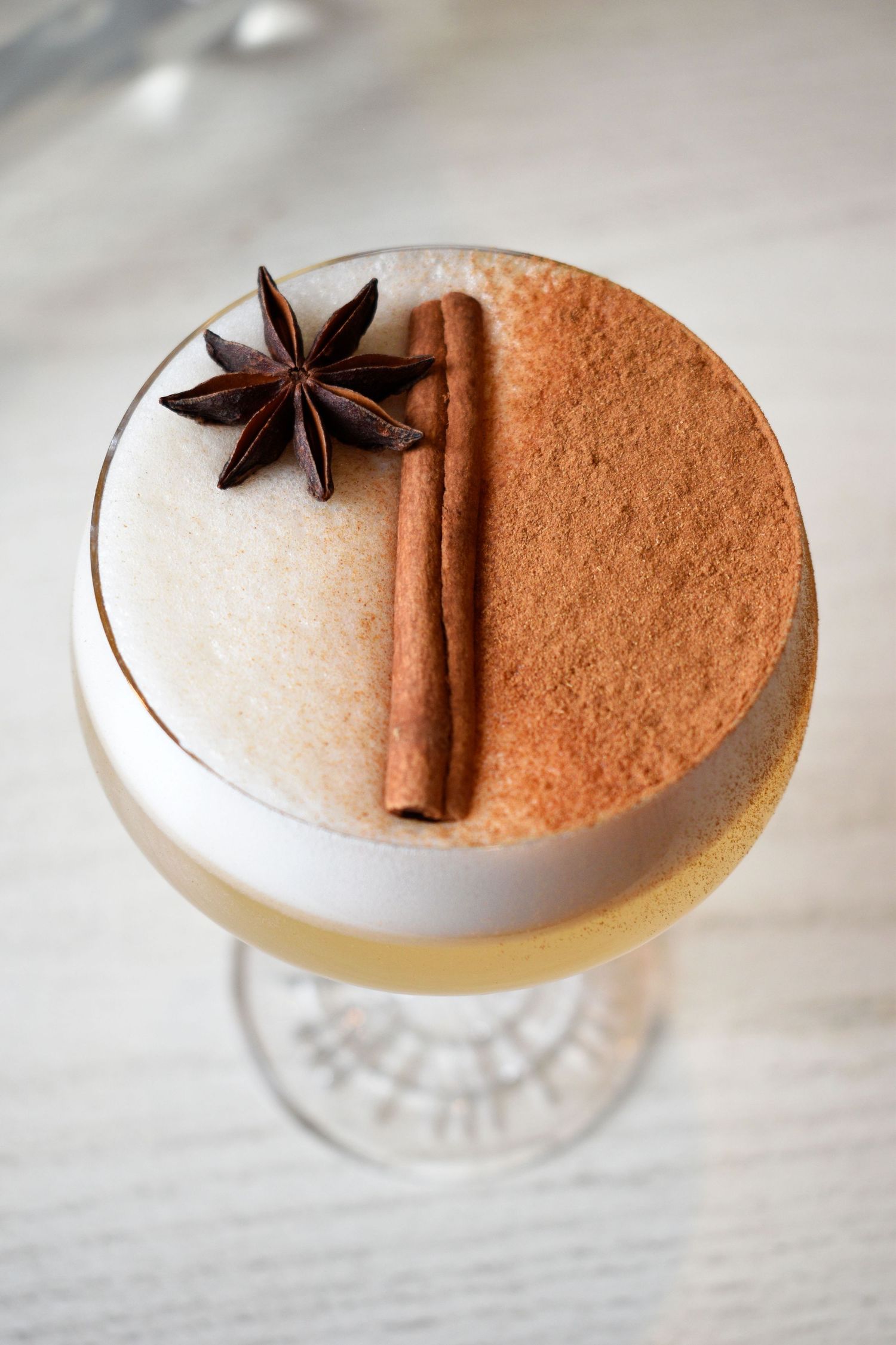 A creamy cocktail topped with foam, dusted cinnamon, a cinnamon stick, and star anise garnish in a coupe glass.
