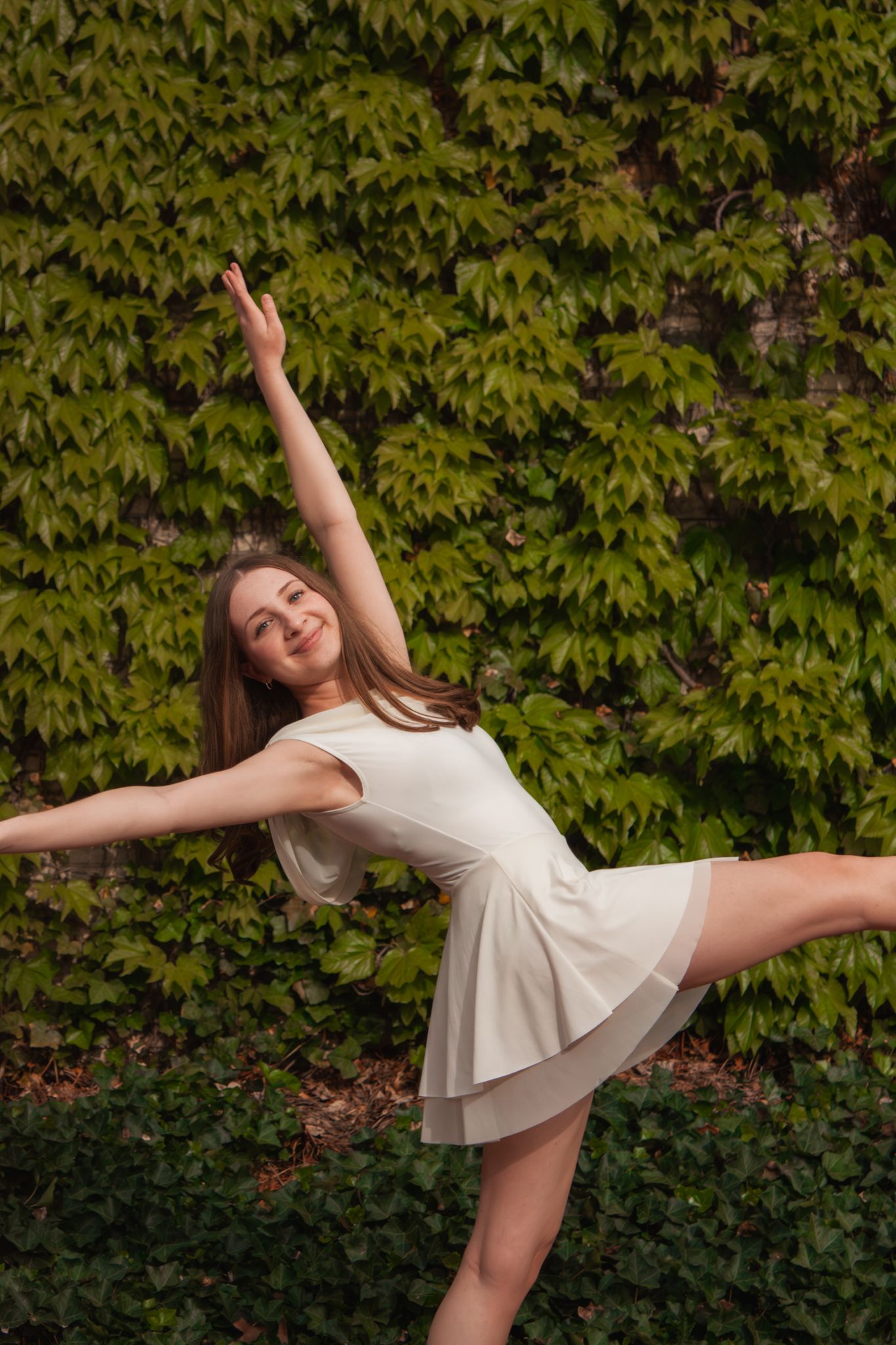 Young dancer in white dress performs elegant one-legged balance pose in front of green ivy wall.