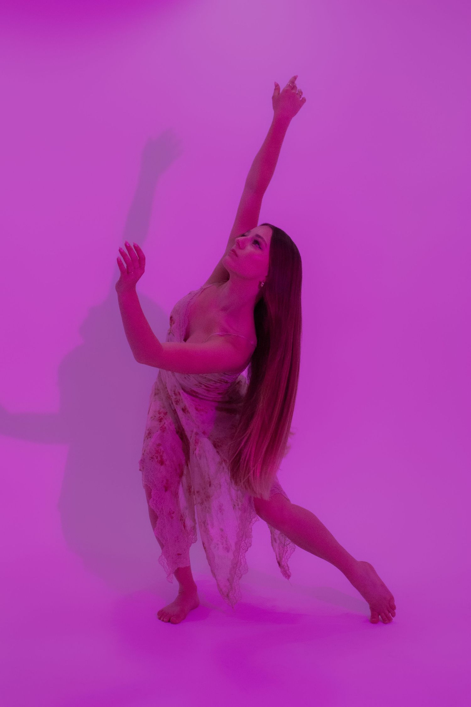 Dancer in flowing dress performs expressive pose with raised arm against vibrant pink studio background.
