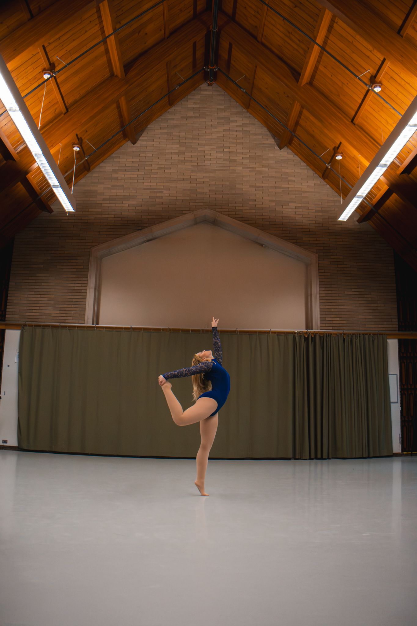 Blonde dancer in blue leotard performs a standing leg hold in a large wooden-ceiling dance hall.