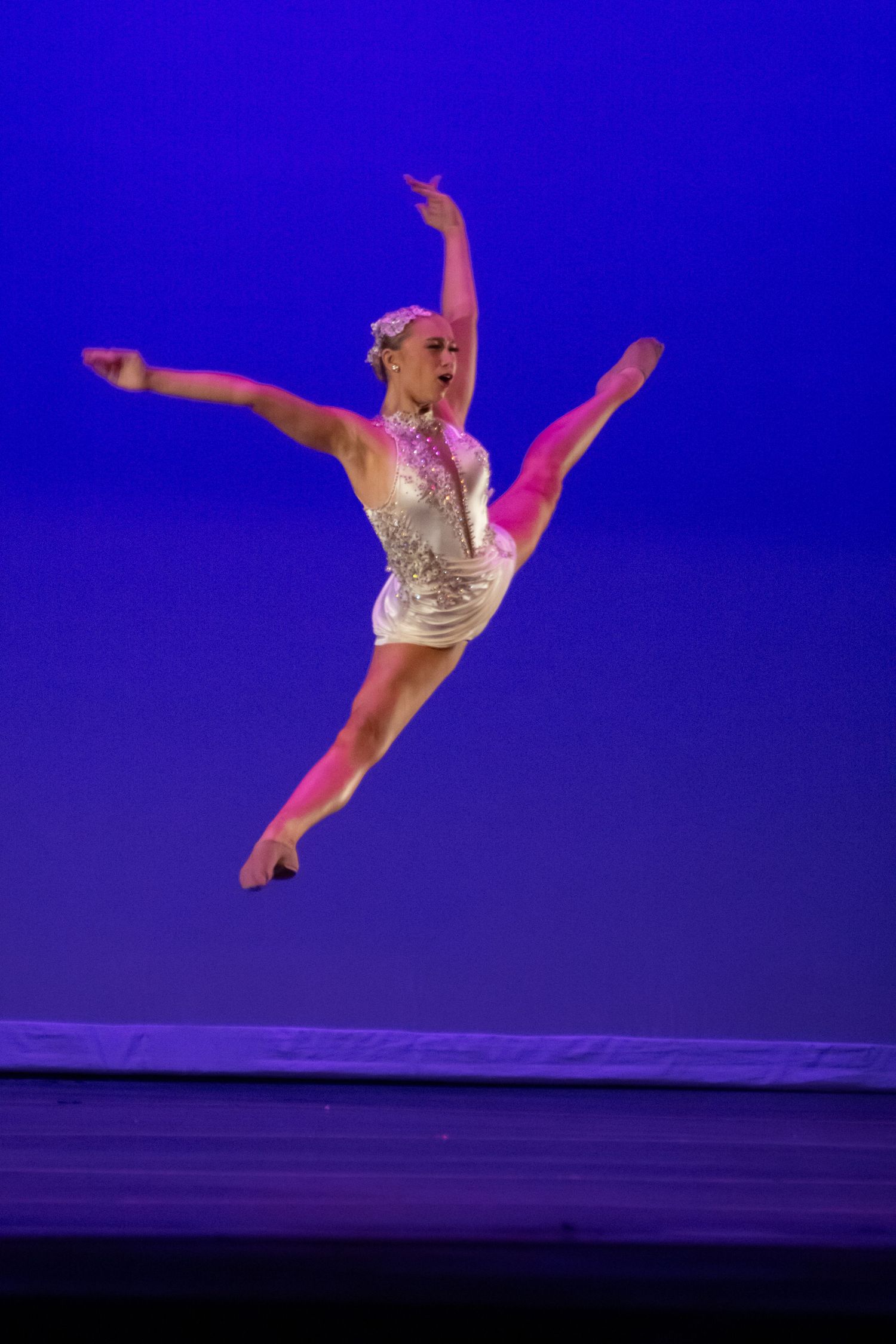 A blonde dancer in a silver sequined leotard performs a grand jete leap on a blue-lit stage.