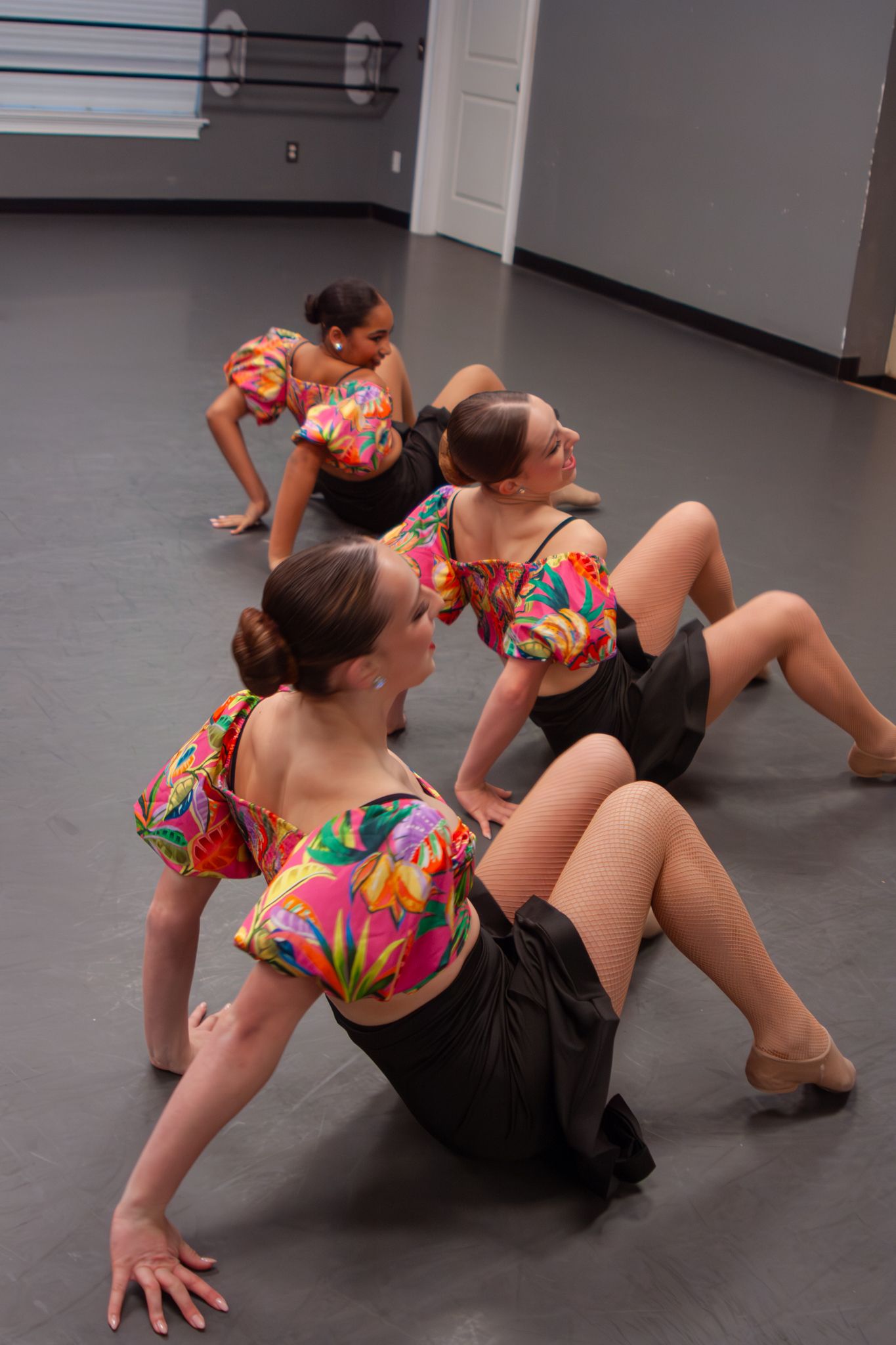 Four dancers in colorful floral tops and black skirts perform a synchronized floor routine in a dance studio.